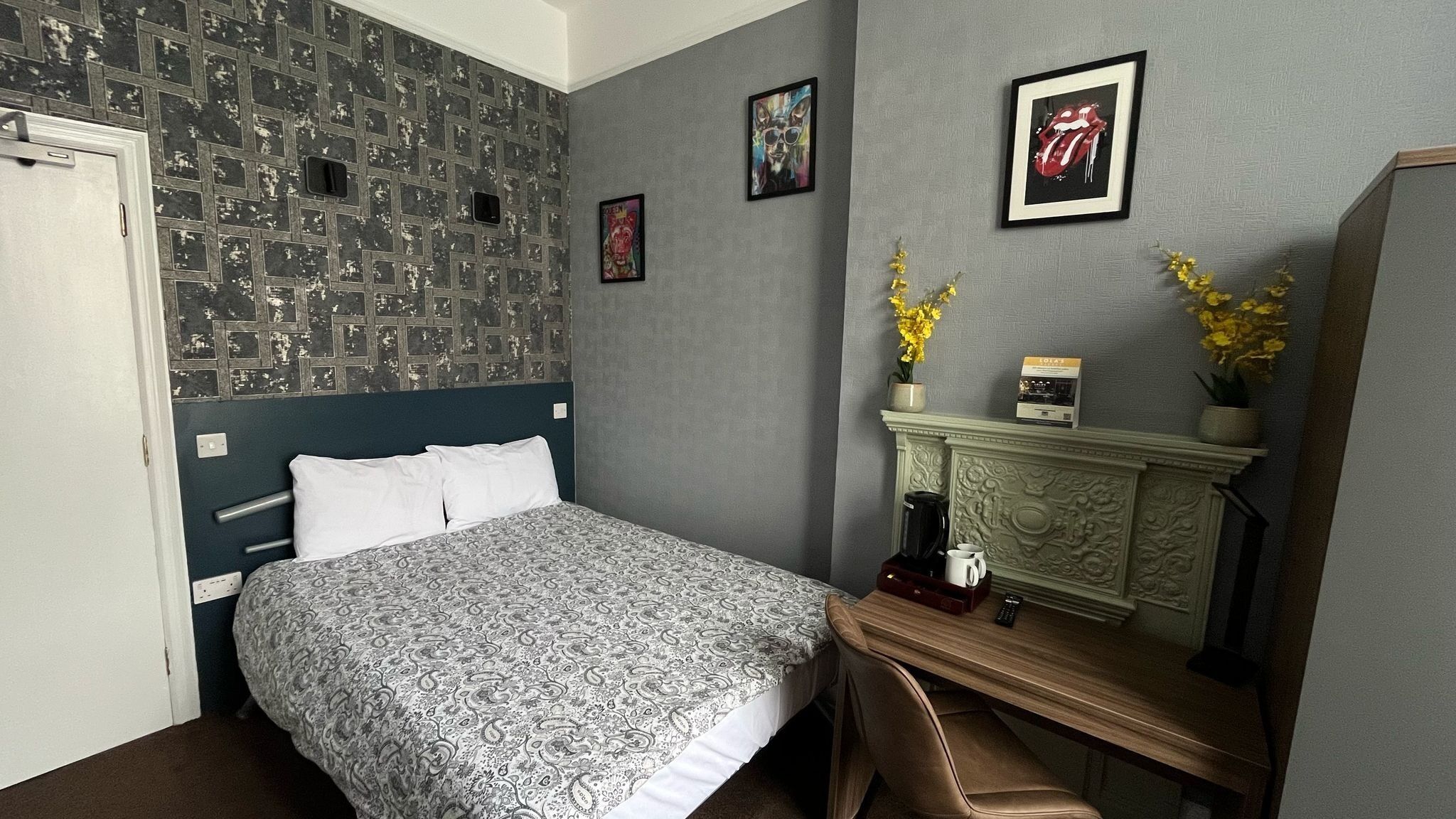 A hotel room with a patterned wall, a bed with a patterned bedspread, a desk with a coffee maker, a mirror, and framed pictures on the wall.