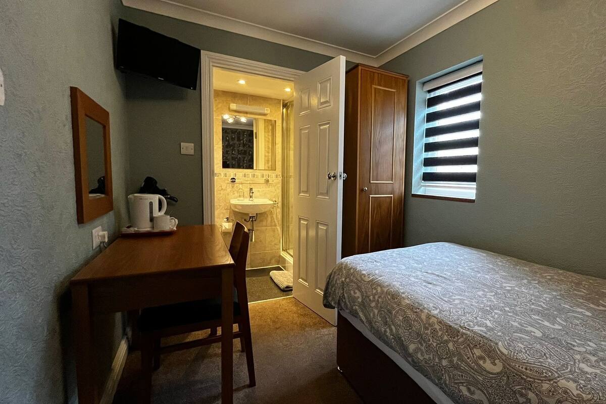 A small bedroom with a bed, a desk, a chair, a window with blinds, and a bathroom visible through an open door.