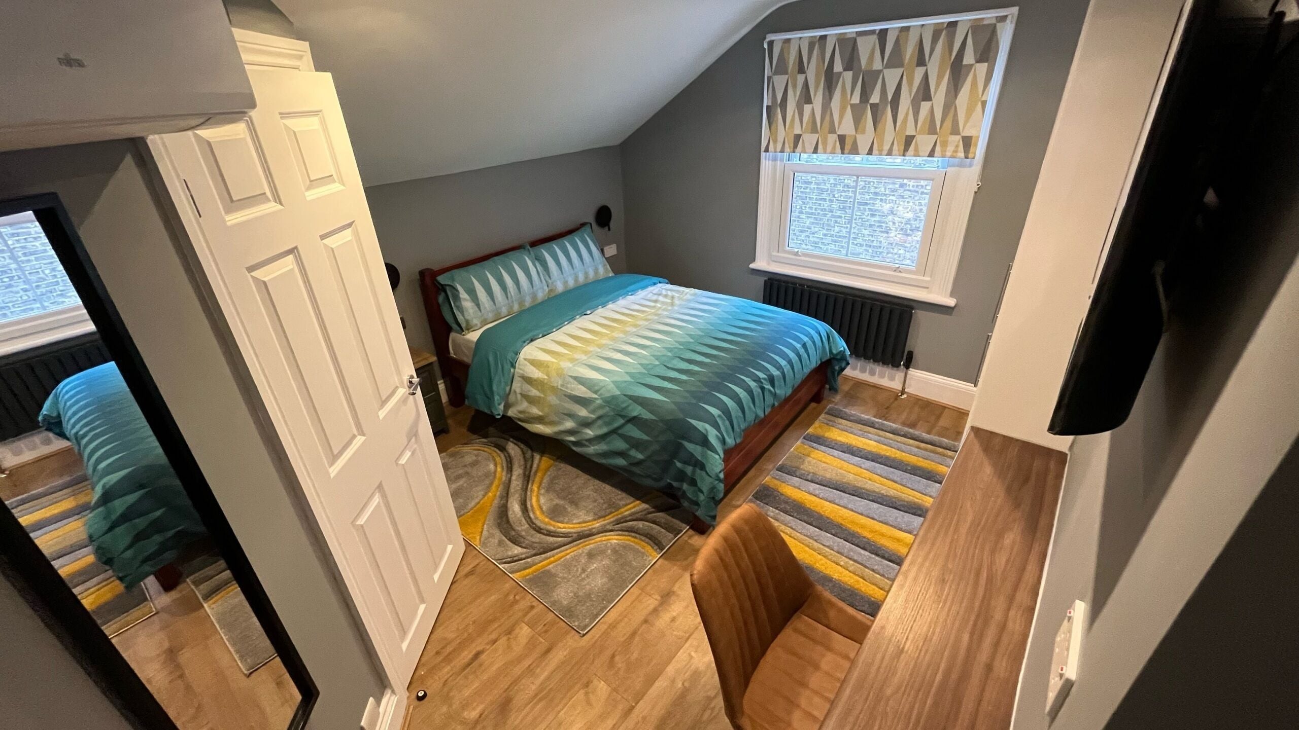 A bedroom with a bed, a window with geometric blinds, a radiator, and a wooden floor.