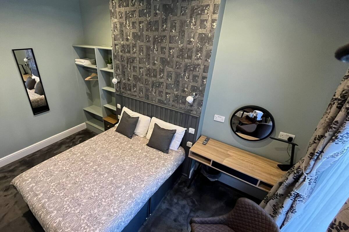 A bedroom with a bed, a mirror, a round mirror, a shelf with folded towels, and a patterned wall.