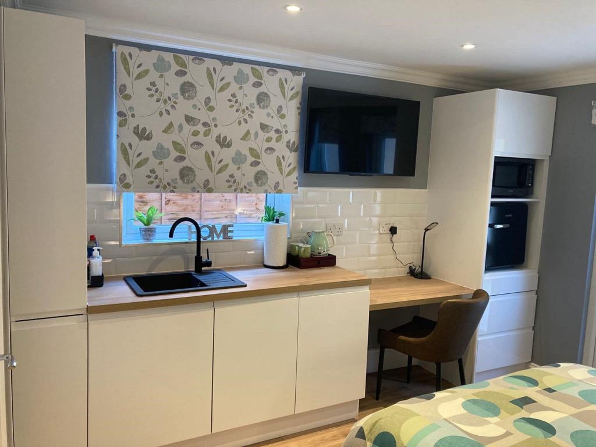A compact kitchen with a sink, a window with a floral curtain, a microwave, and a small chair.