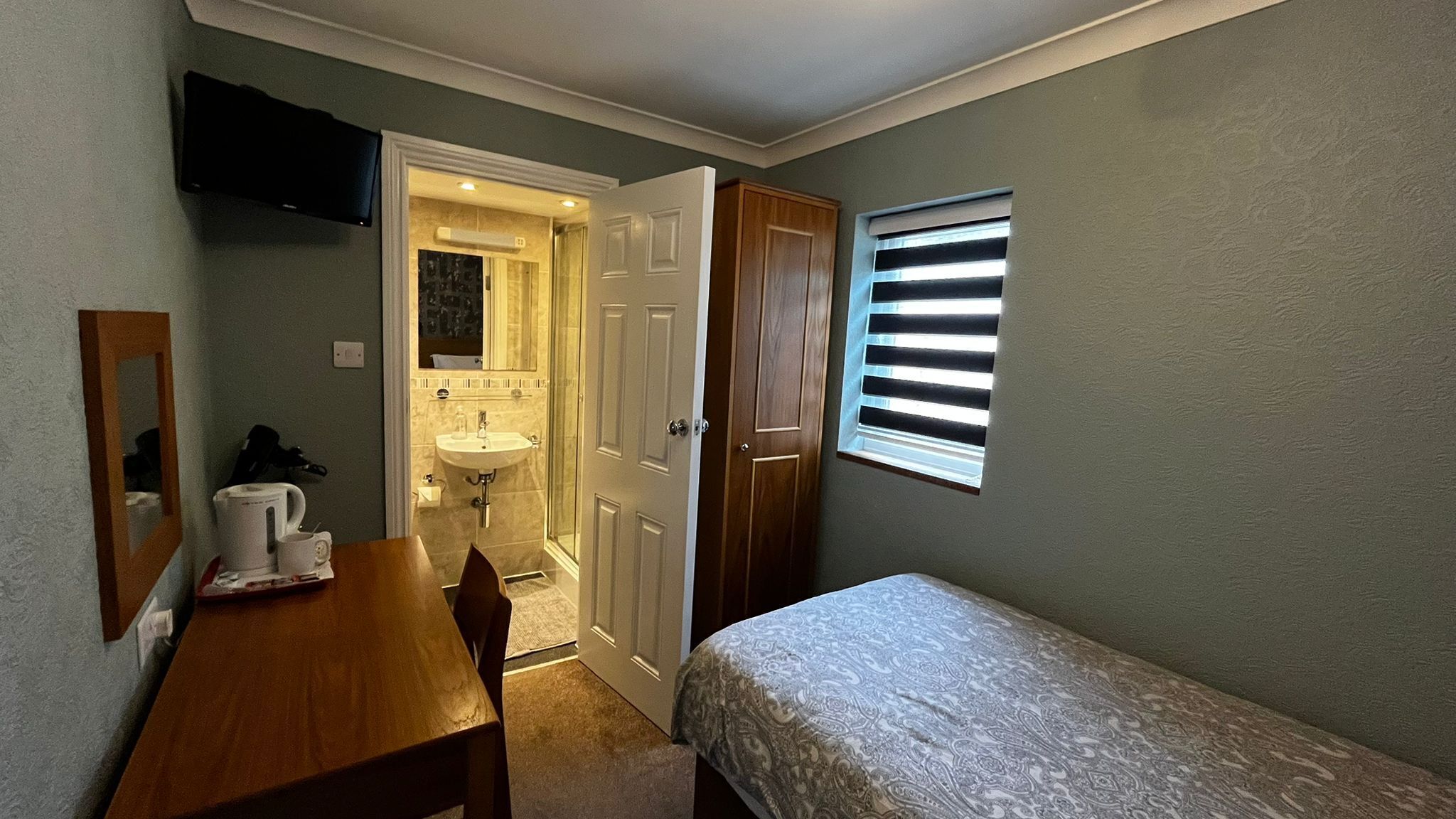 Una habitación pequeña con una cama individual, una mesita con una silla, un lavabo y una ventana con persianas.