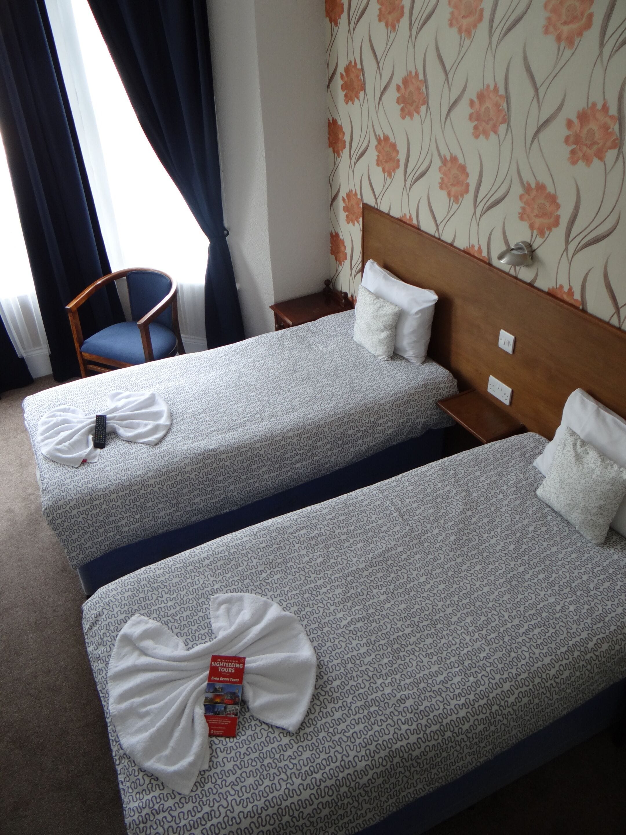 A hotel room with two single beds, a wooden headboard, a small bedside table, a chair, and a window with curtains.