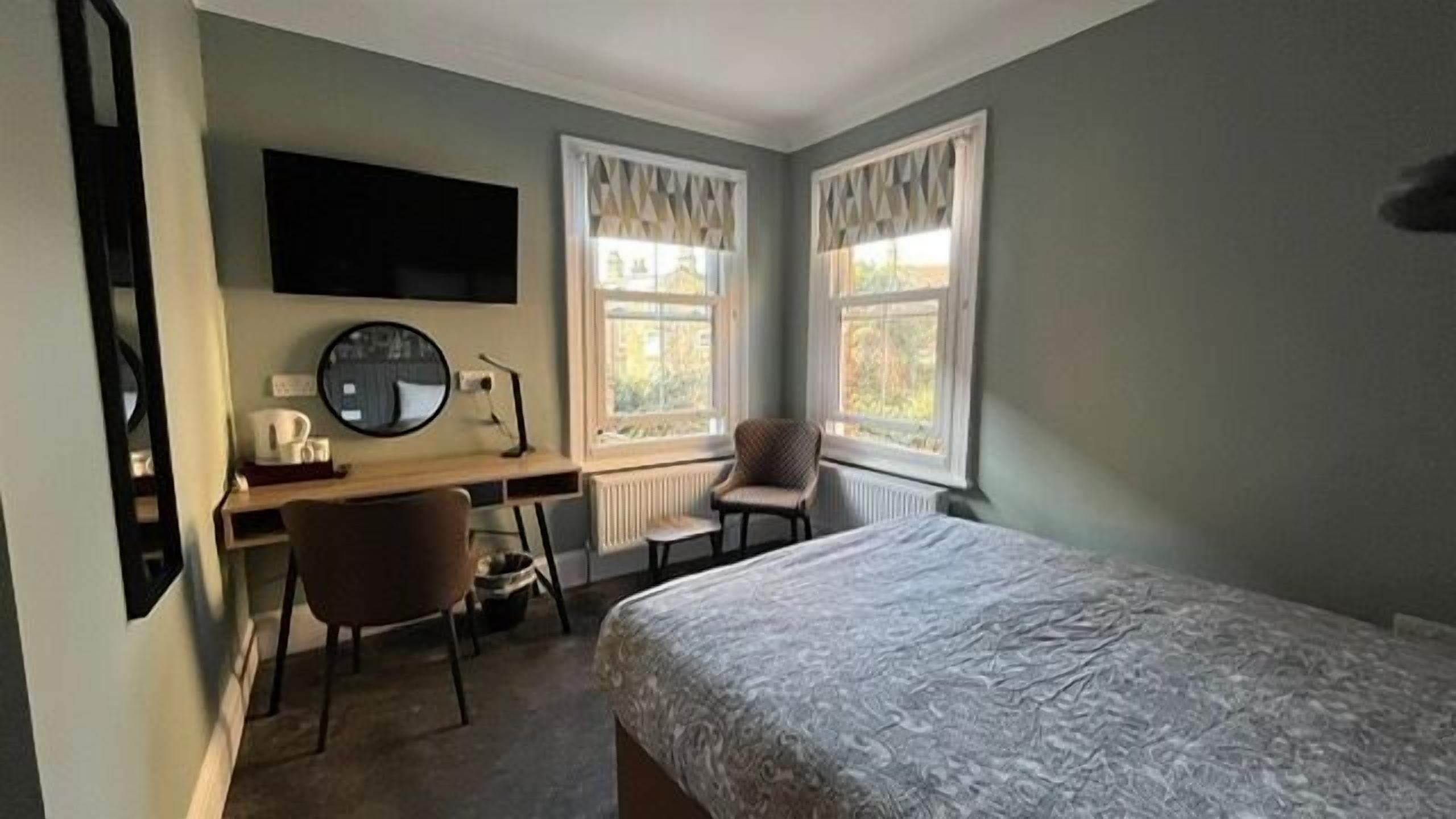 A bedroom with a bed, a desk with a chair, a mirror, and two windows with curtains.