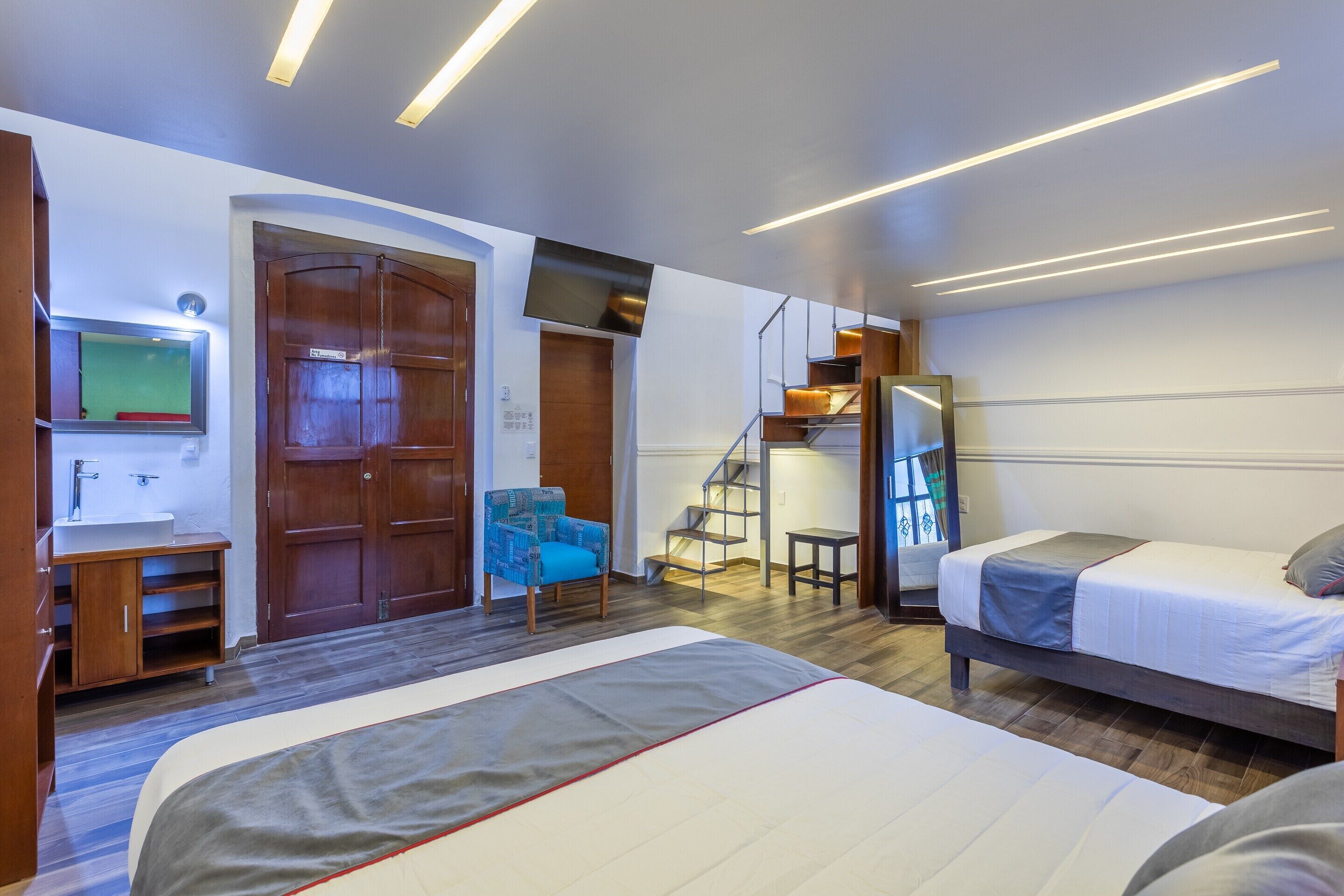 A modern hotel room with a large bed, a chair, a desk with a mirror, a wardrobe, and a staircase.