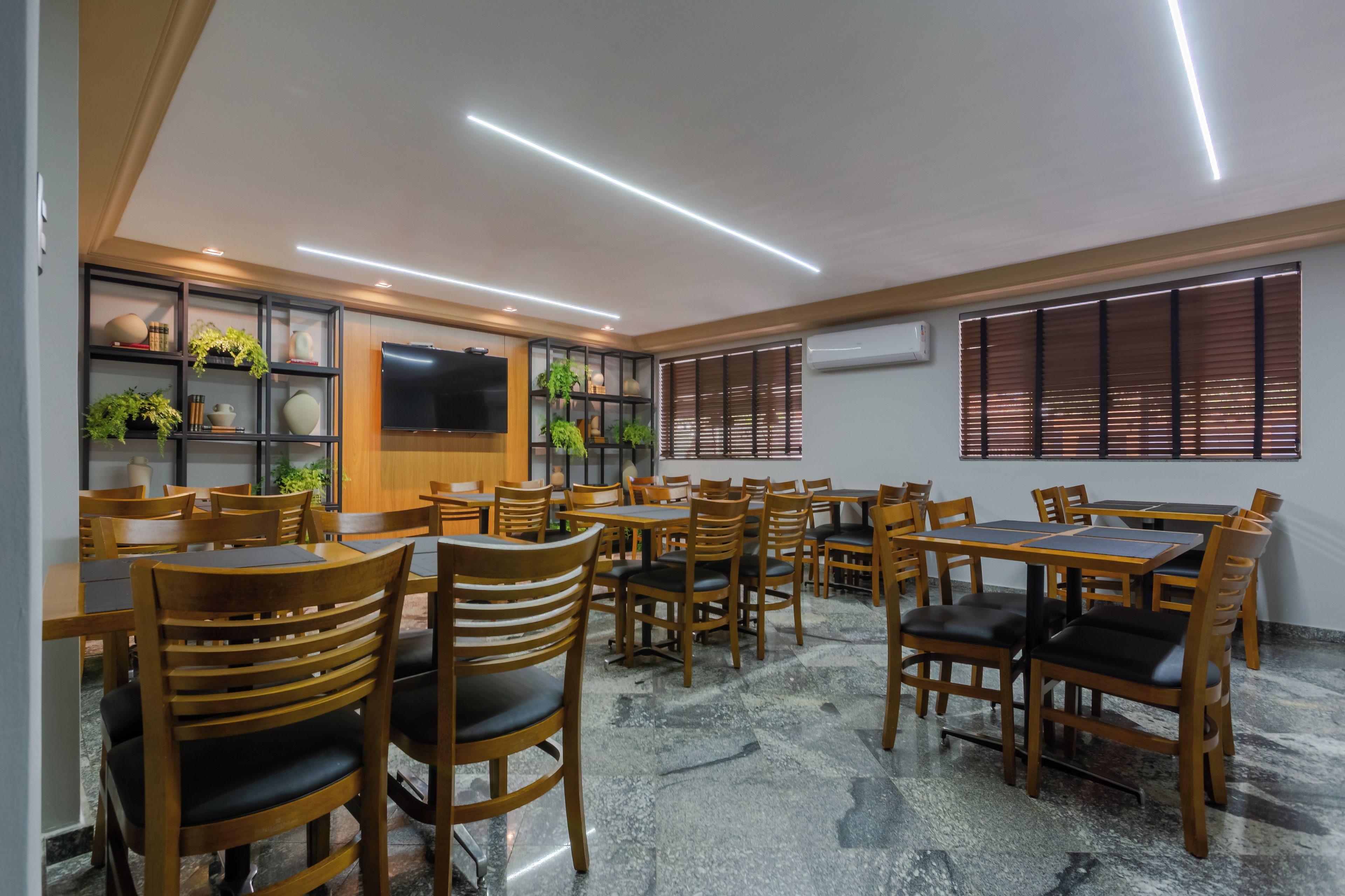 A restaurant with multiple tables and chairs, a flat-screen TV mounted on the wall, and shelves with plants and decorative items.