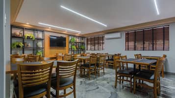 A restaurant with multiple tables and chairs, a flat-screen TV mounted on the wall, and shelves with plants and decorative items.