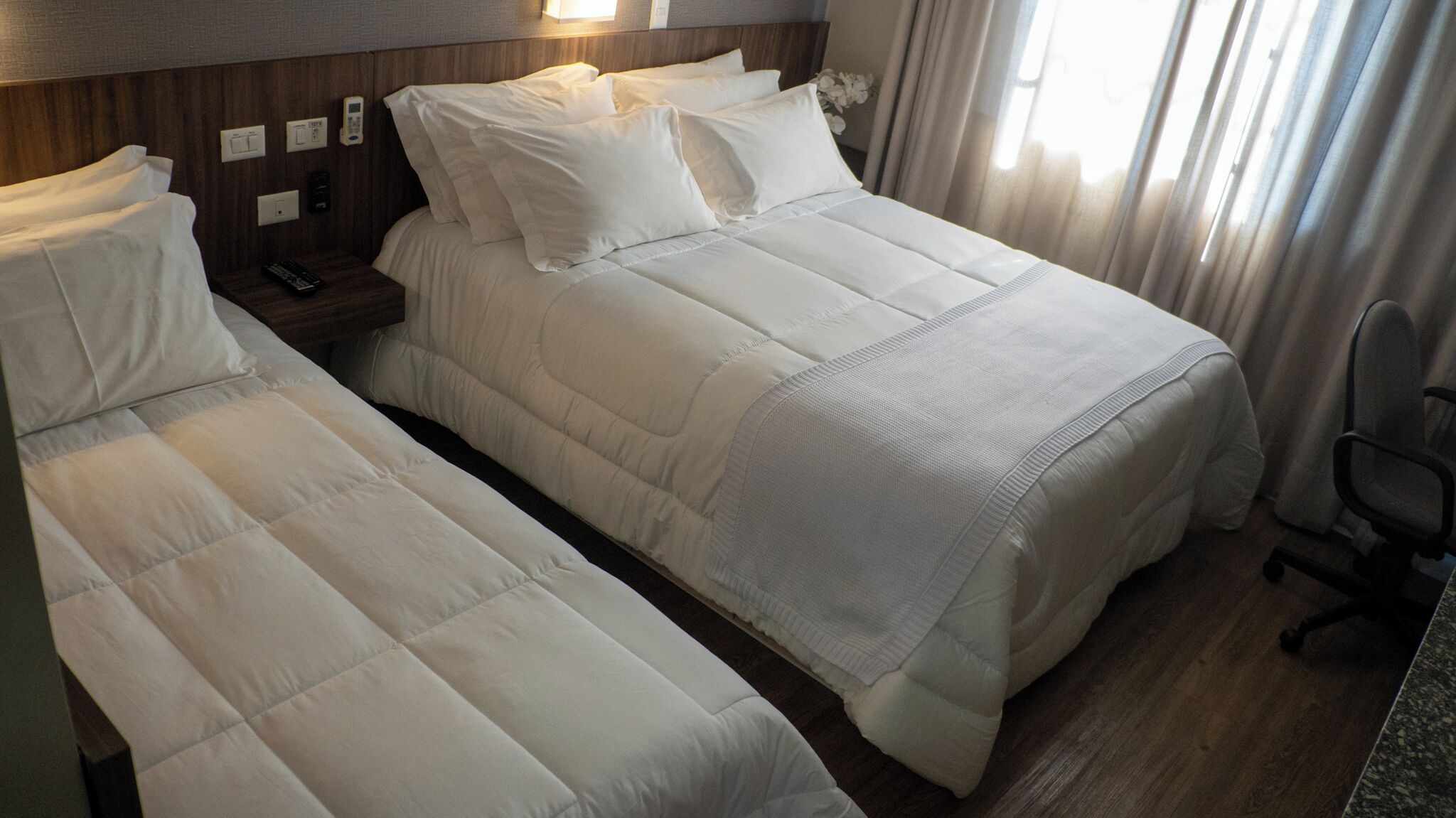 Habitación de hotel con dos camas, una mesita de noche, una silla y un ventanal con cortinas.