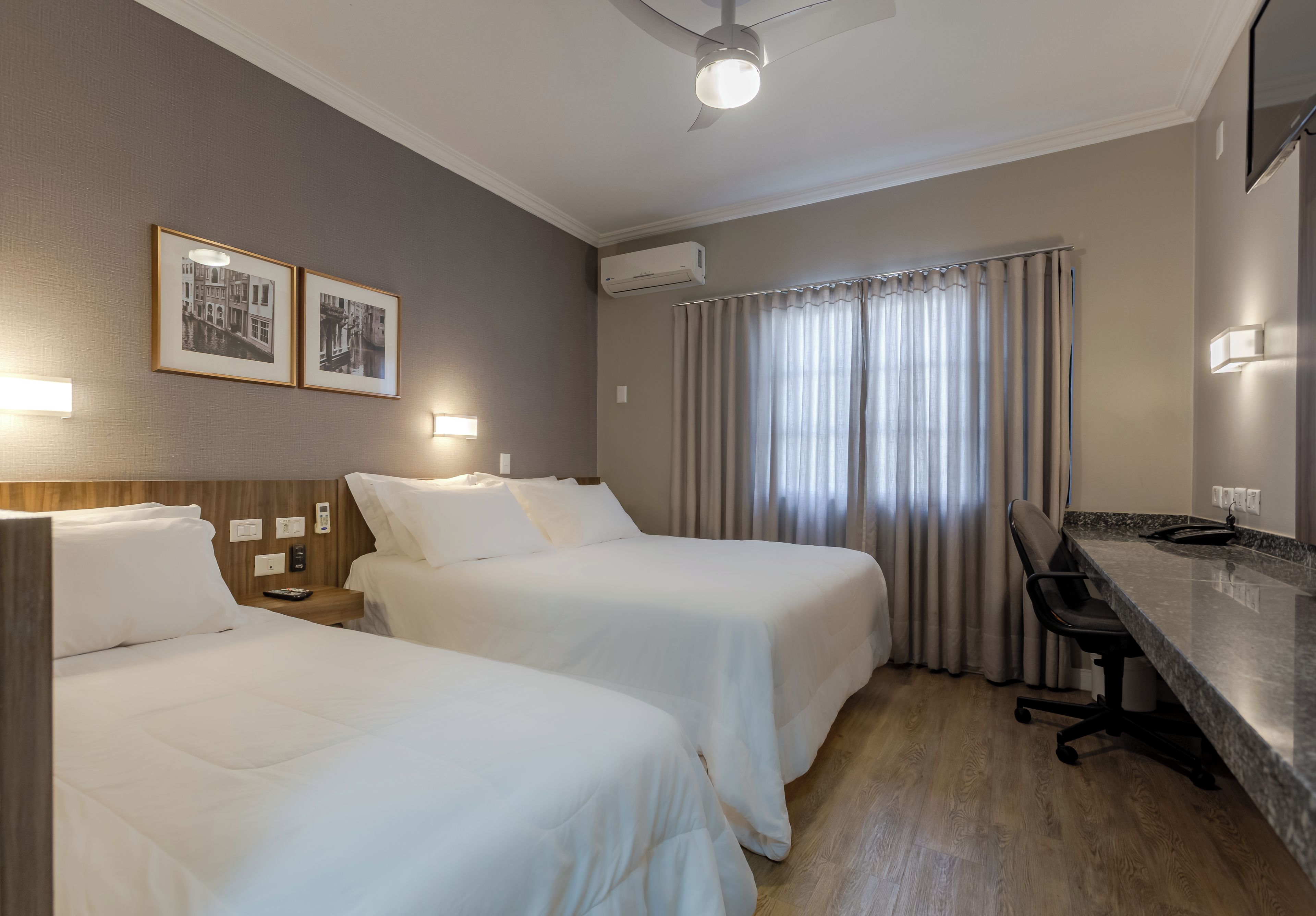 Habitación de hotel con dos camas, un escritorio, una silla, un ventilador de techo y dos cuadros en la pared.