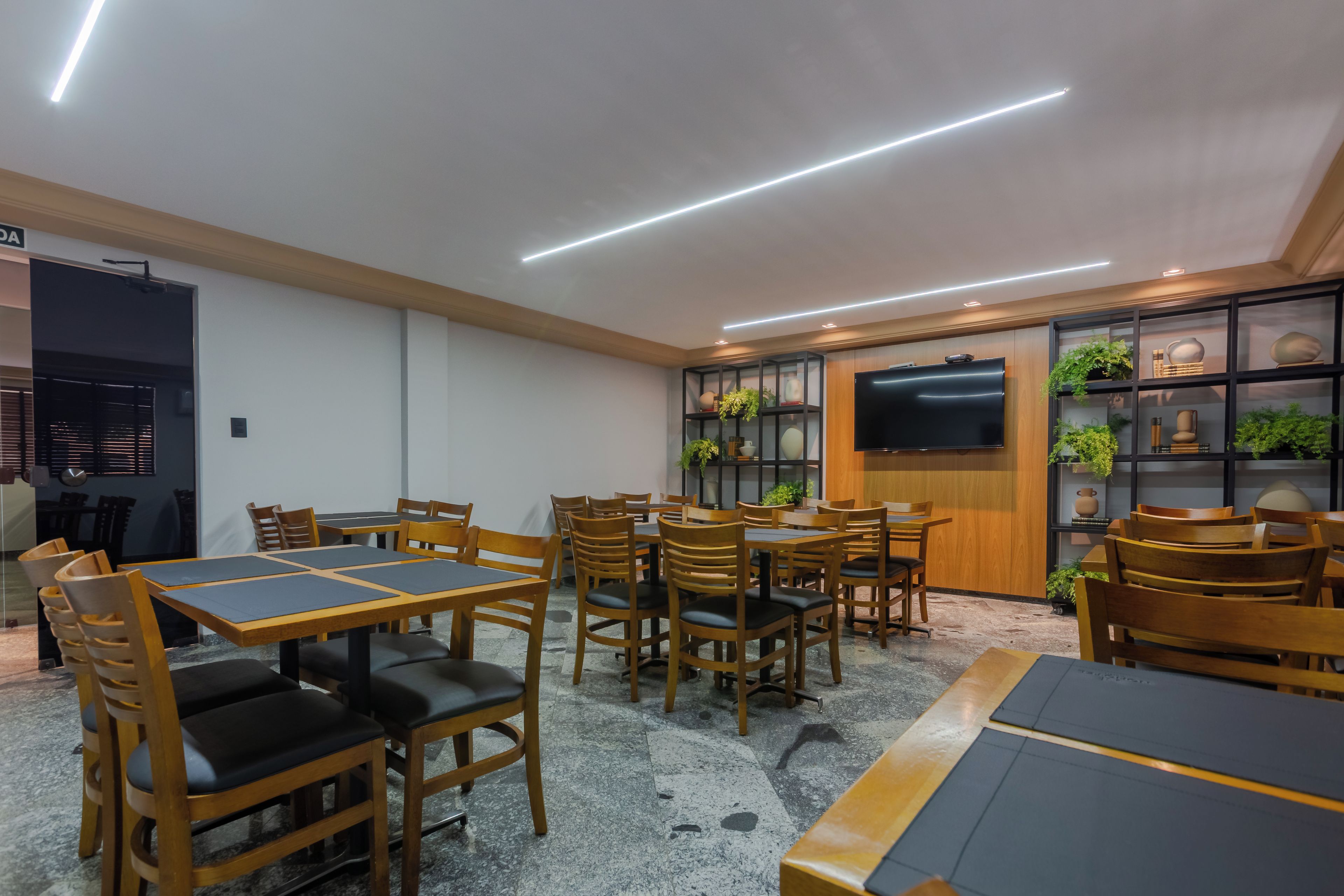 Un intérieur moderne avec des tables et des chaises en bois, un téléviseur à écran plat fixé au mur et des étagères ornées d’objets décoratifs.