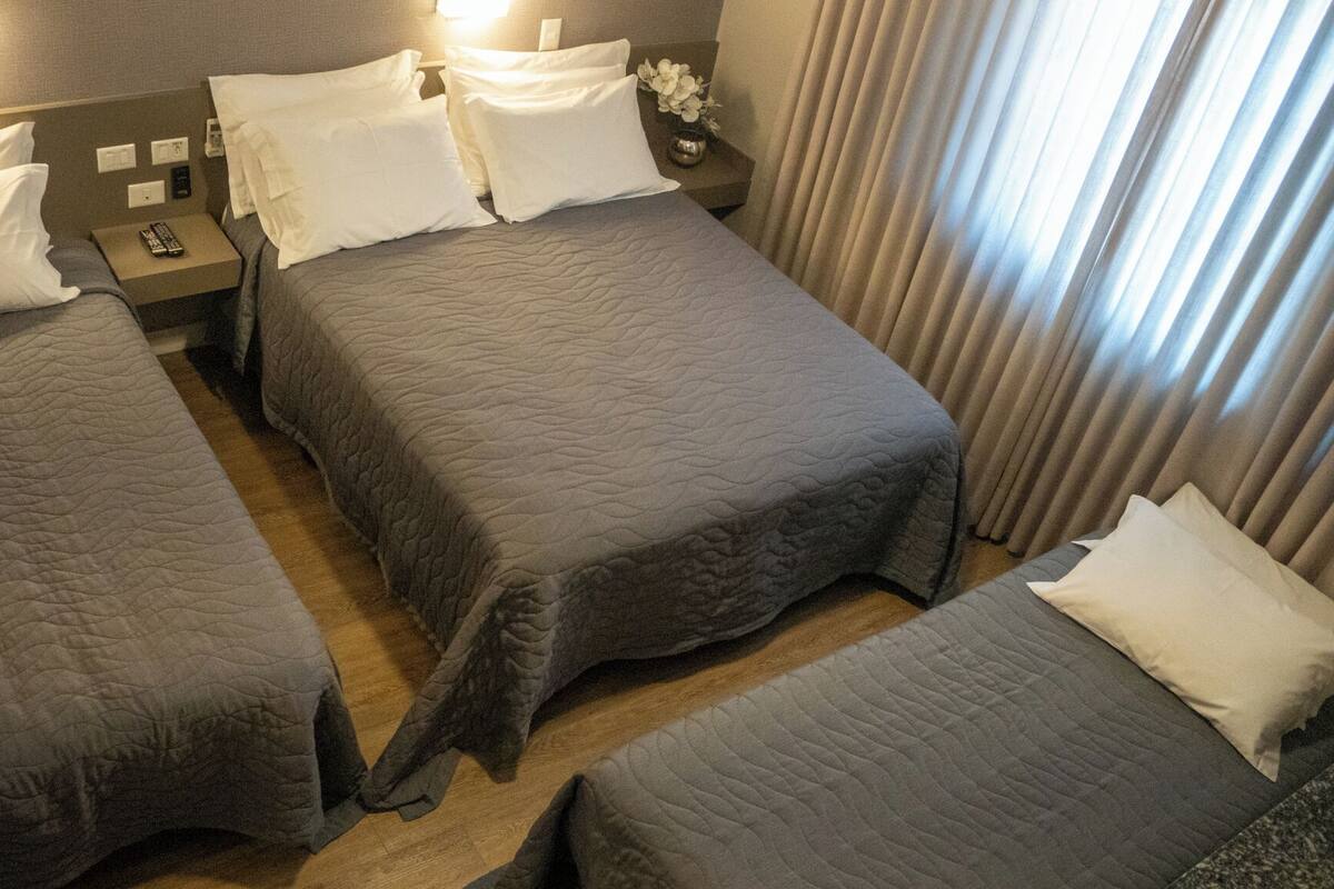 Una habitación de hotel con tres camas, una mesita de noche, una lámpara y una ventana con cortinas.