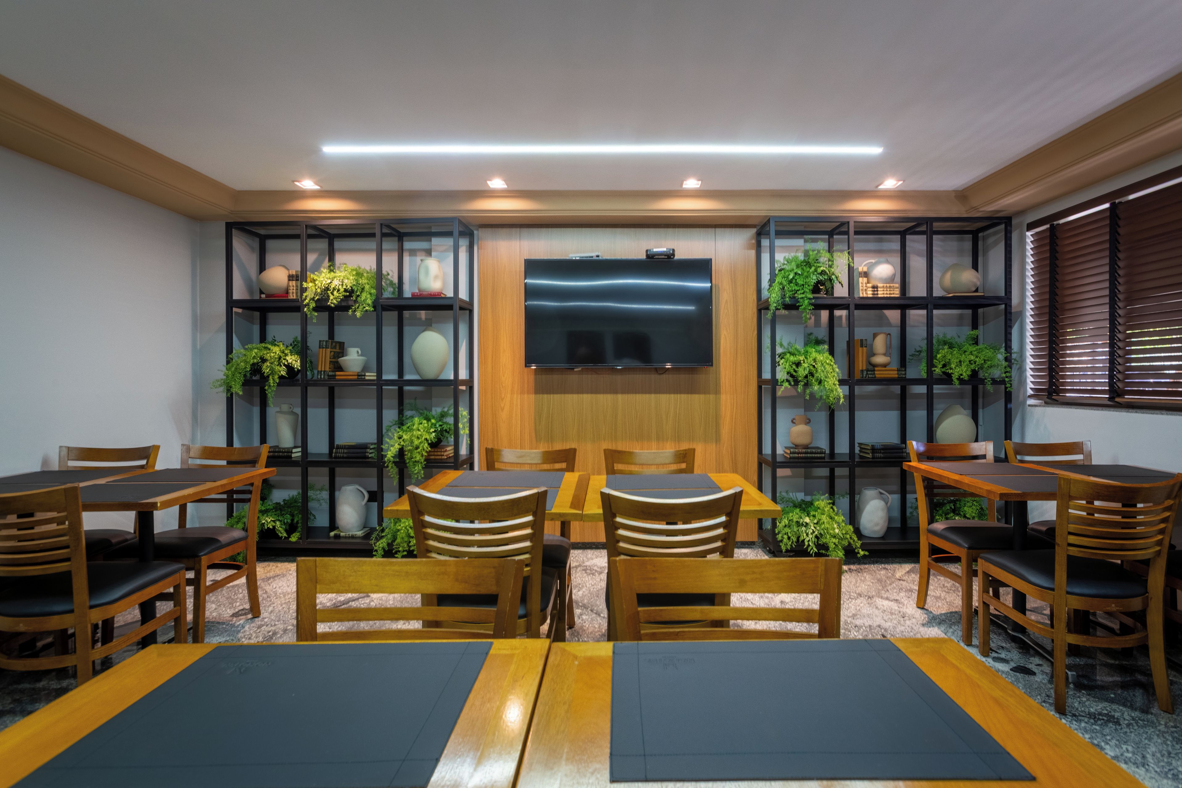 A conference room with a large flat-screen TV, wooden tables, and chairs, surrounded by shelves with plants and decorative items.