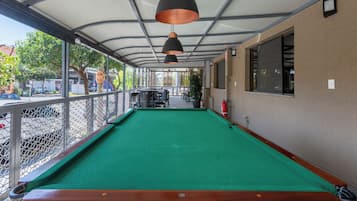 Uma mesa de bilhar com superfície verde aveludada, rodeada por uma grade de metal, sob uma área coberta com teto de metal e iluminação pendente.