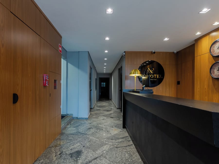 A hotel reception area with a wooden reception desk, a black clock, and a sign that reads 'NO HOTEL'.