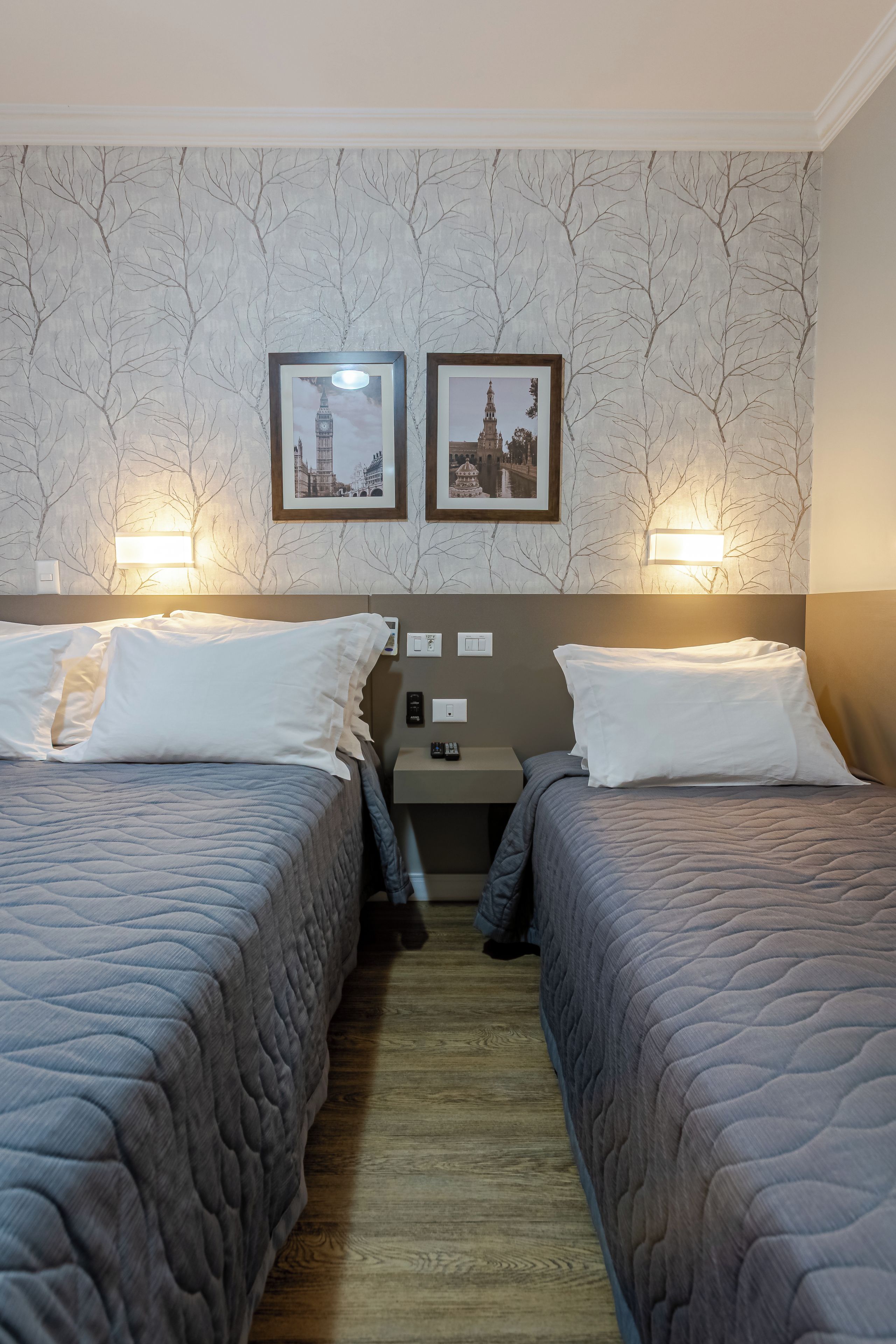 Habitación de hotel con dos camas individuales, una mesita de noche, una lámpara de pared y dos cuadros enmarcados.
