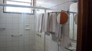 Un baño con azulejos blancos, cabina de ducha con mampara de cristal, toallero con toallas blancas, espejo y secador de manos.