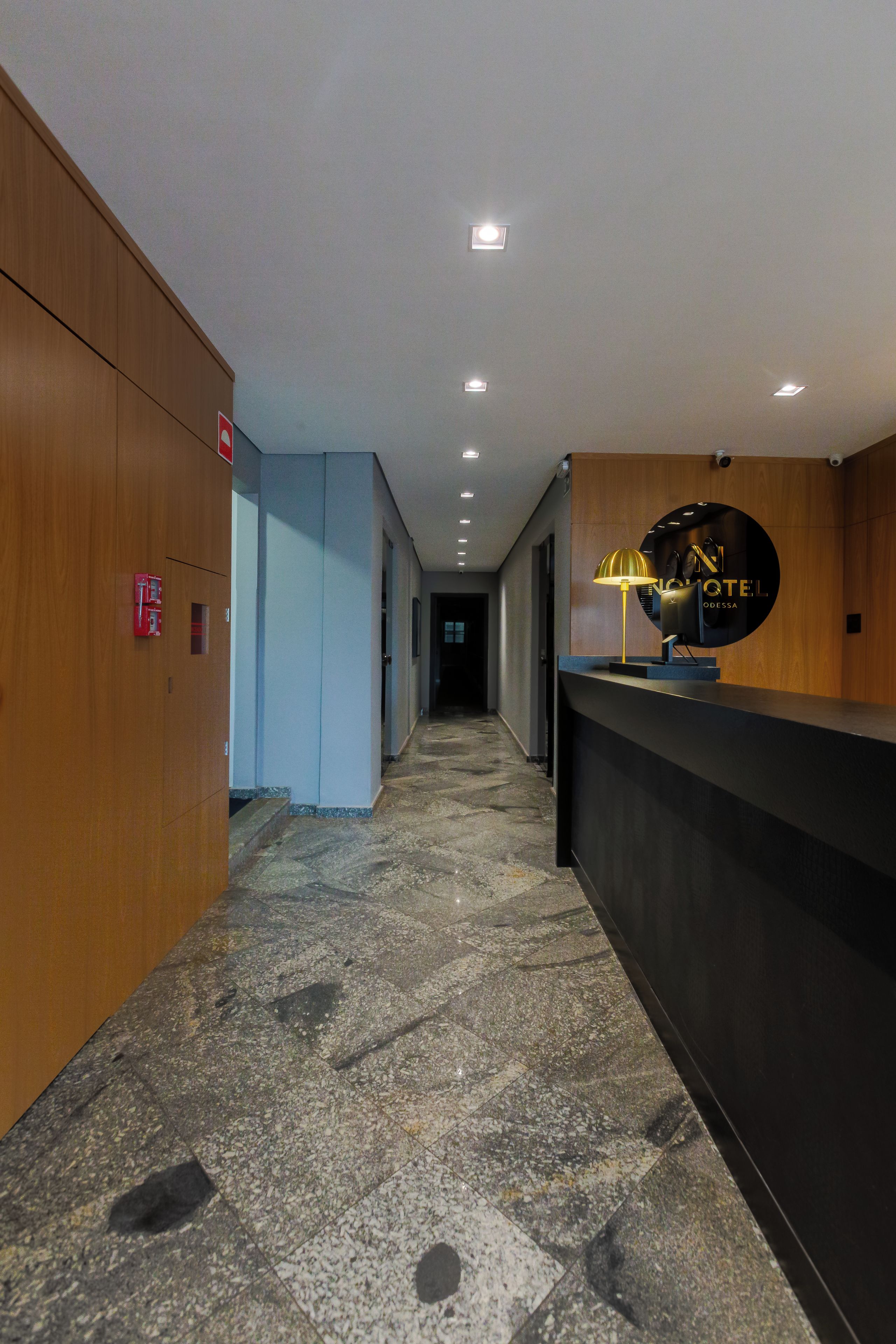Zona de recepción de un hotel con mostrador, un reloj en la pared y un detector de incendios a la izquierda.