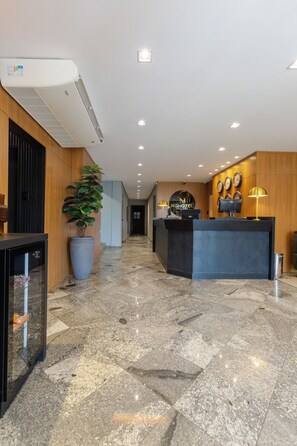 Zona de recepción de hotel con mostrador, una planta en maceta, un reloj de pared y una pantalla digital.