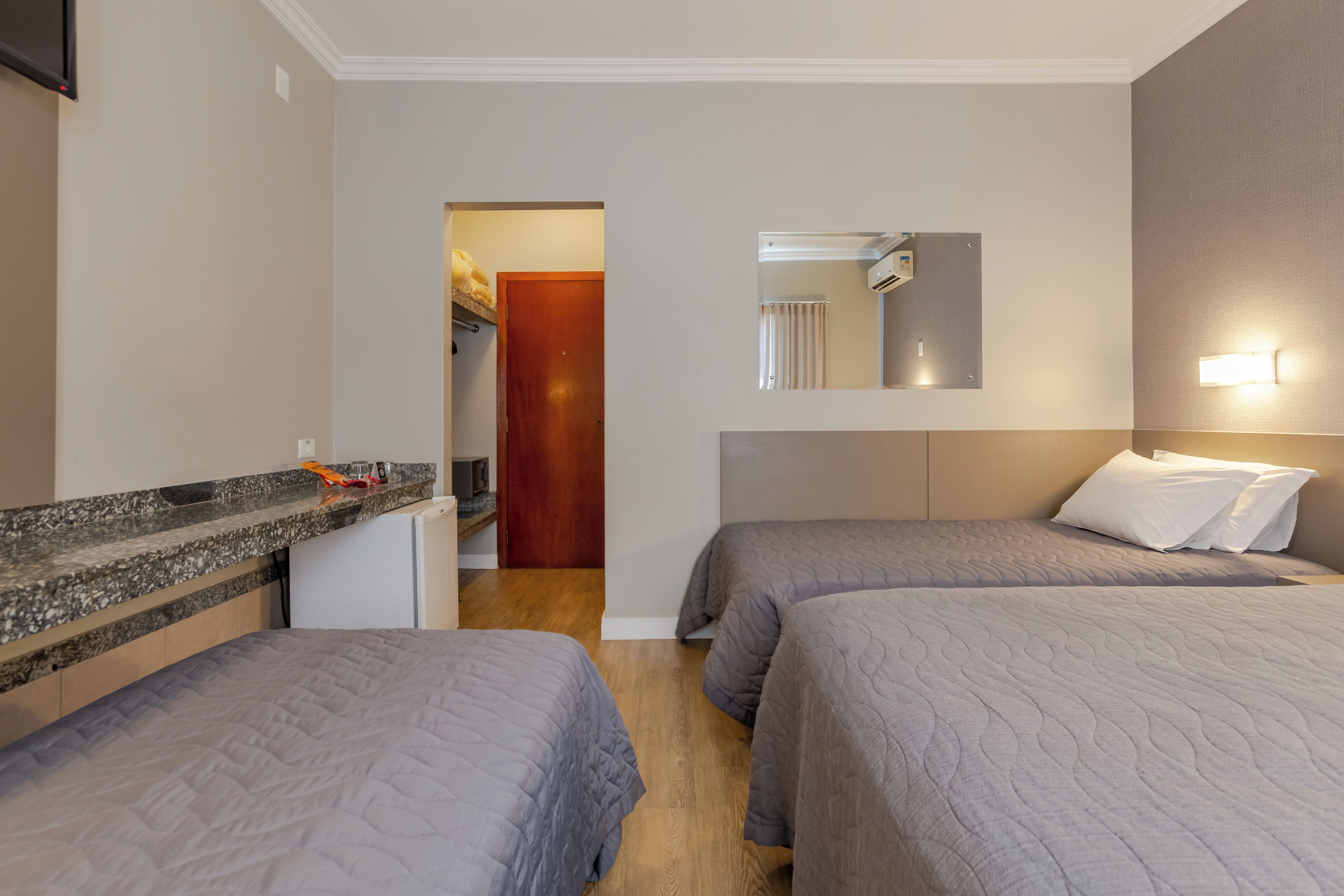A hotel room with two beds, a small kitchenette, a mirror, and a door leading to another room.