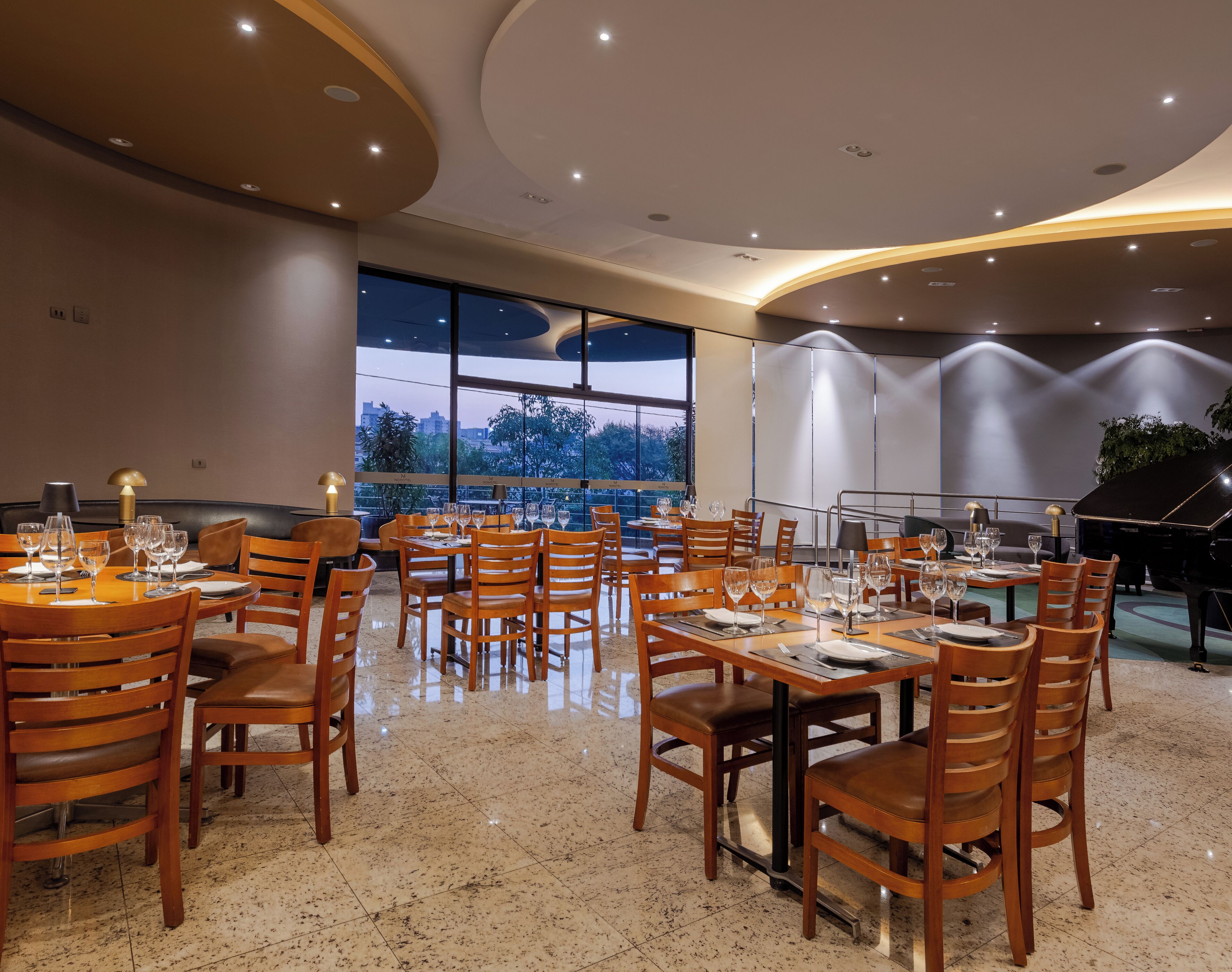 A restaurant with multiple tables and chairs, large windows, and a piano.
