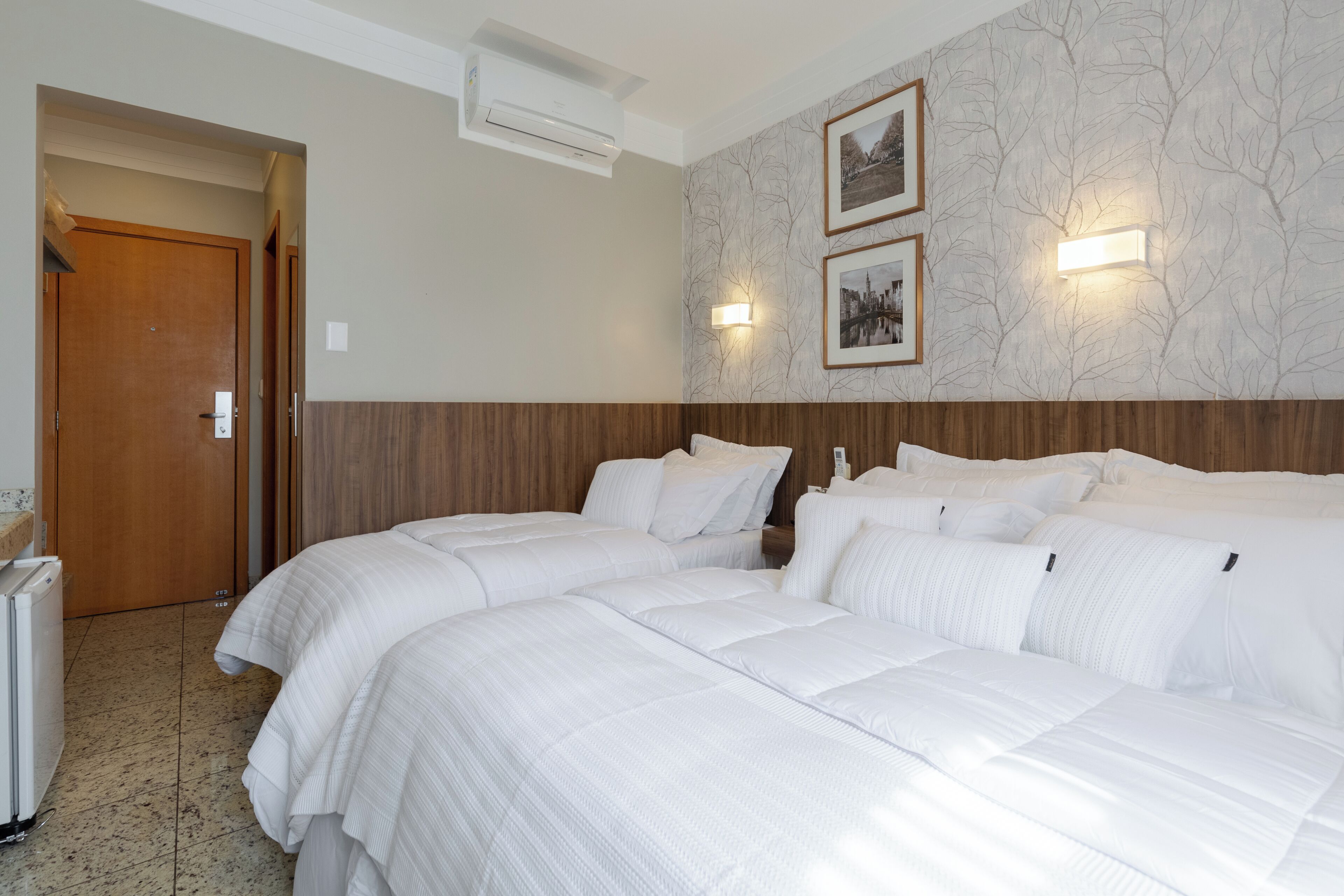 Habitació d'hotel amb dos llits, un panell de capçalera de fusta, aire condicionat fixat a la paret i dos quadres emmarcades.