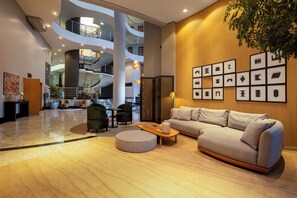 Un moderno vestíbulo de hotel con una escalera curva, un área de descanso con un sofá y sillones, y una pared decorada con obras de arte enmarcadas.
