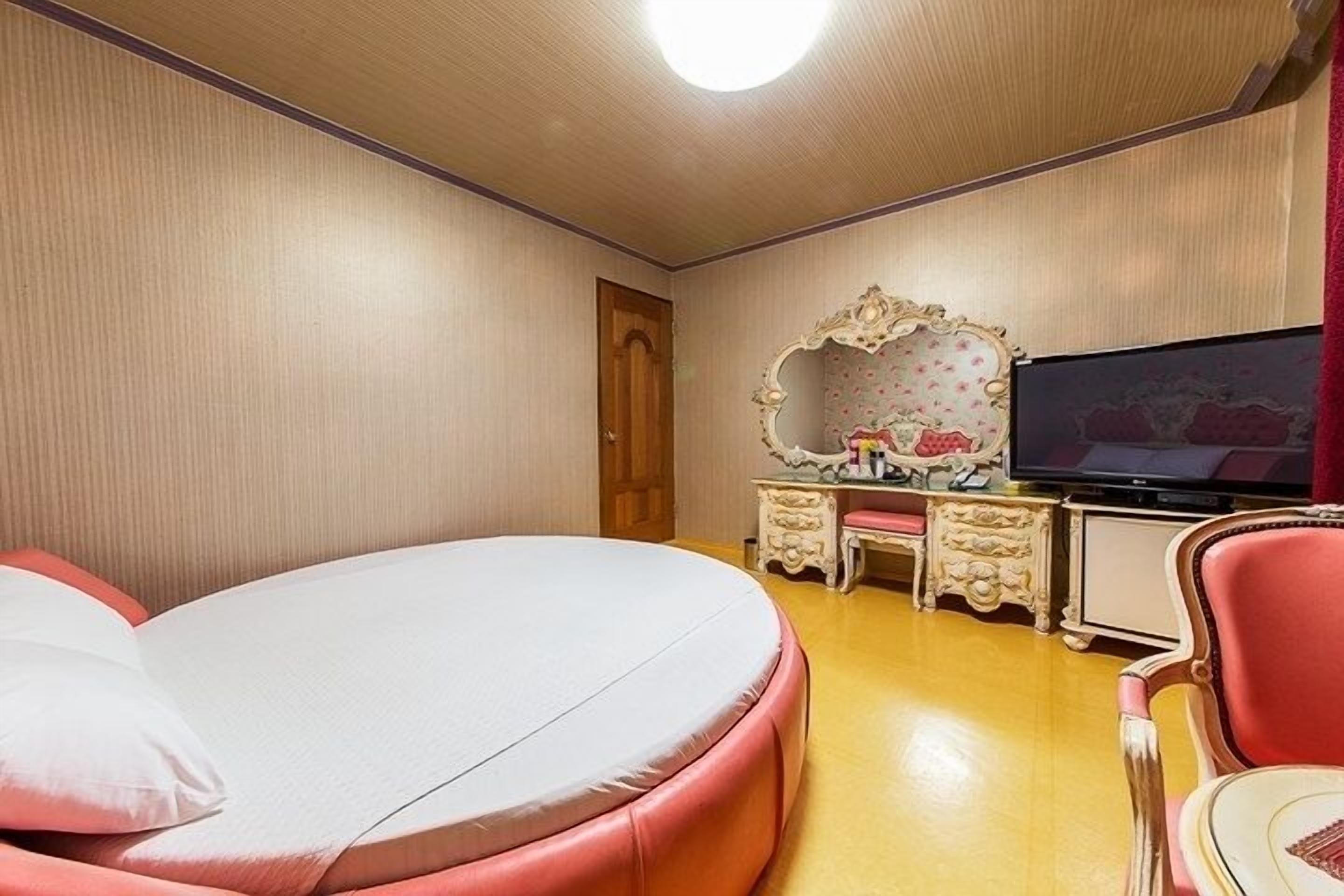 A room with a round bed, a television, a vanity with a mirror, and a red chair.