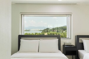 Habitación de hotel con cama, mesita de noche, control remoto y ventana con vista a un río y montañas.