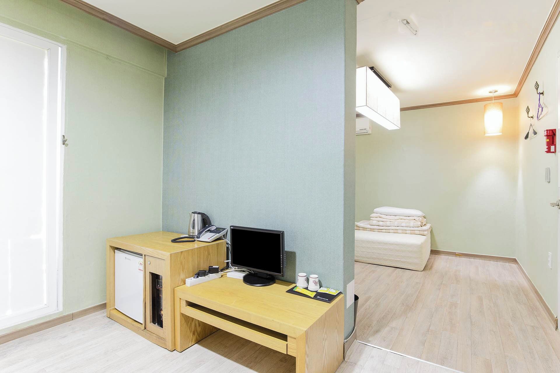 A compact hotel room with a wooden desk, a small refrigerator, a flat-screen TV, a kettle, and a bed with a white comforter.