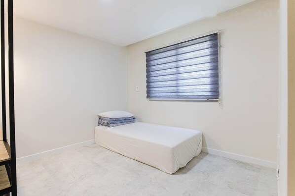 A small, neatly arranged room with a single bed, a bench, and a window with blinds. - La Feliz Hotel CSM (Chuncheon)