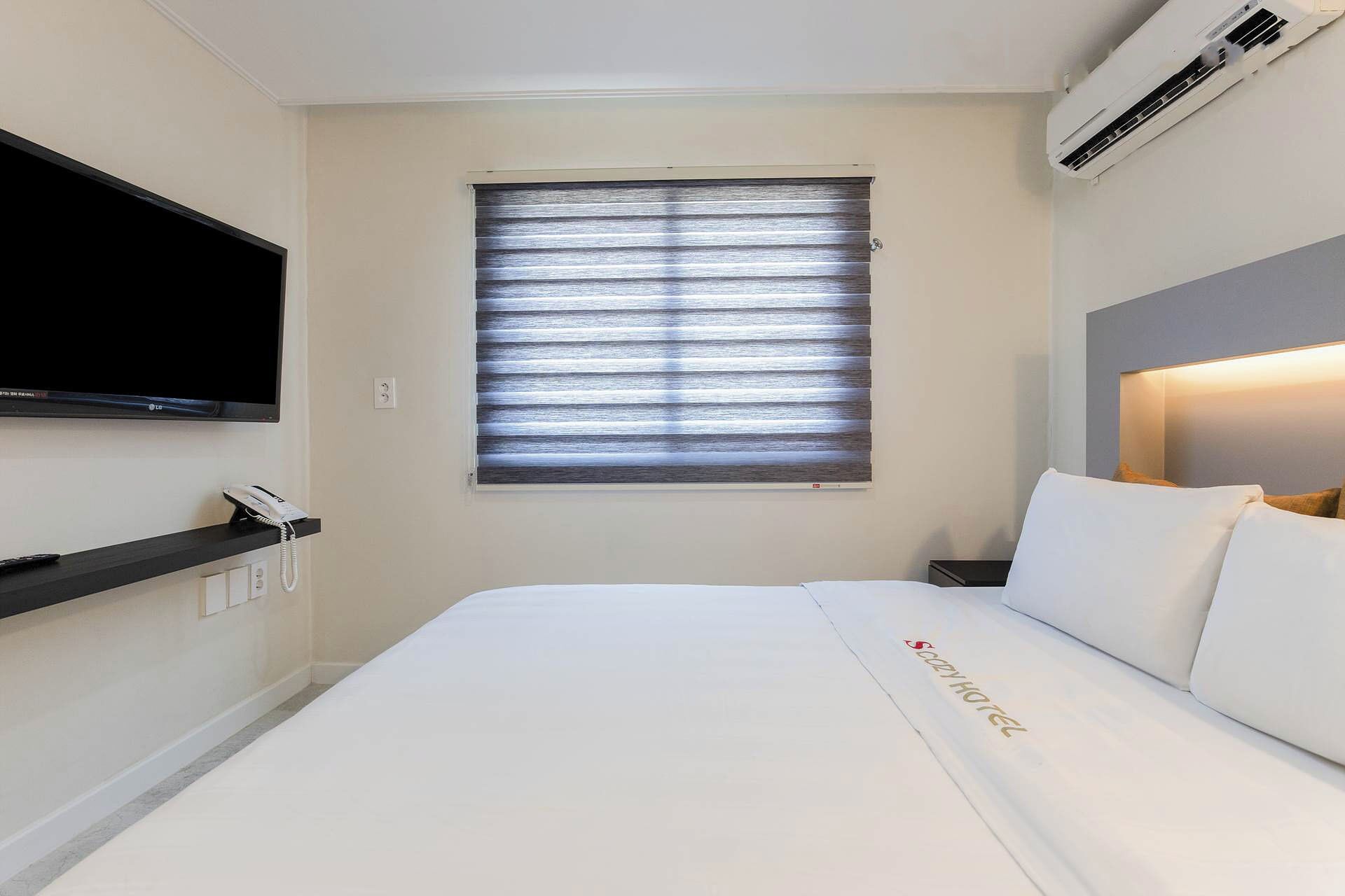 Une chambre d’hôtel comprenant un lit, un téléviseur à écran plat fixé au mur, une fenêtre avec des stores, une table de chevet avec un téléphone et un appareil de climatisation.