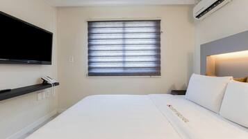 Une chambre d’hôtel comprenant un lit, un téléviseur à écran plat fixé au mur, une fenêtre avec des stores, une table de chevet avec un téléphone et un appareil de climatisation.