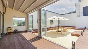 Une vaste terrasse dotée d’une table et de chaises, d’un ventilateur et offrant une vue sur un paysage montagneux grâce à de grandes portes vitrées.