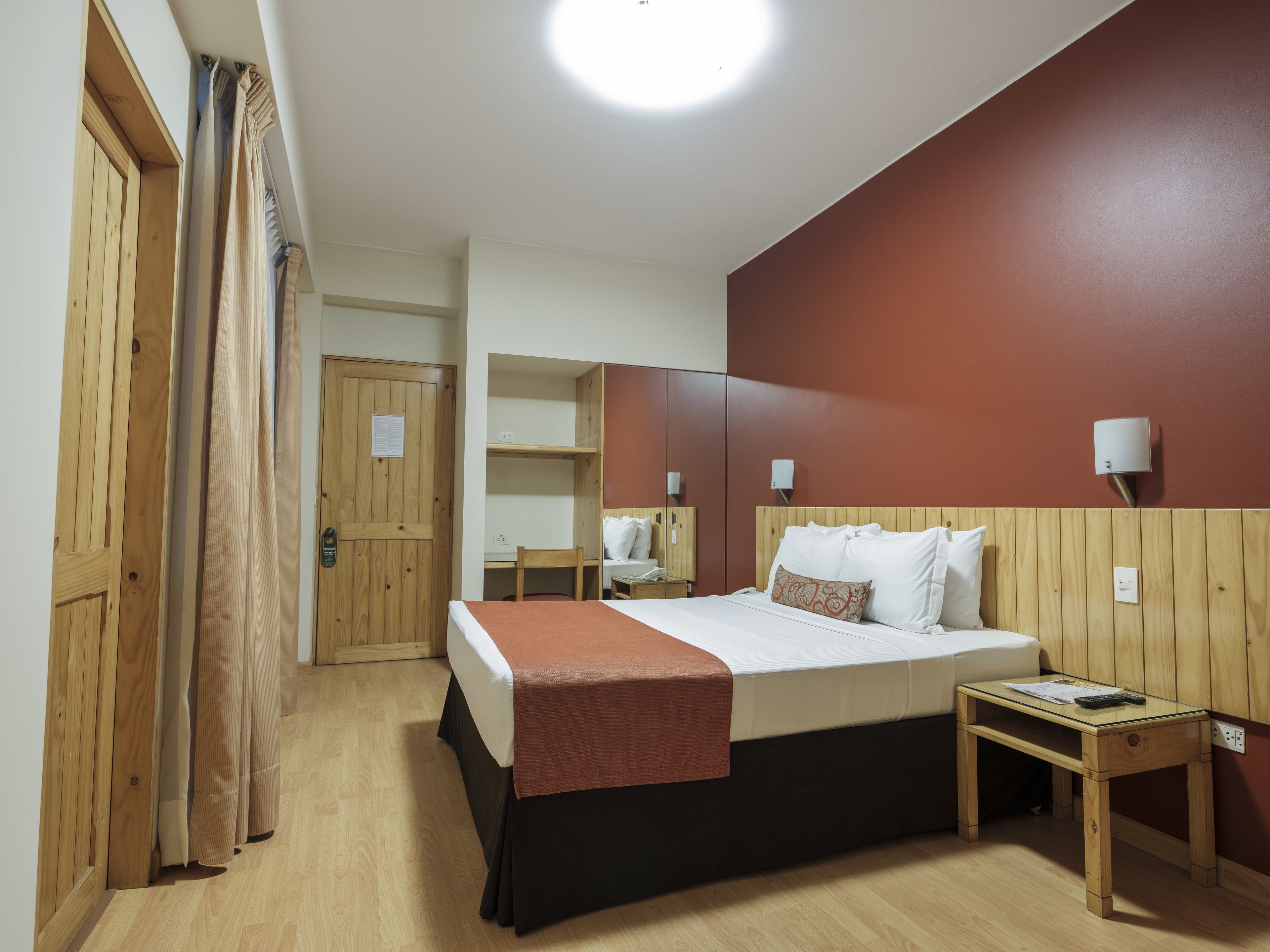 Quarto de hotel com cama, escrivaninha, cadeira, mesa de cabeceira e guarda-roupa.