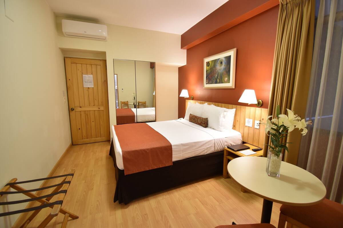 Una habitación de hotel con cama, mesita de noche, lámpara y una pequeña mesa con un jarrón de flores.