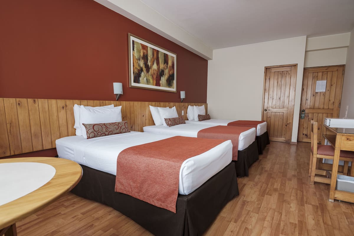 Habitación de hotel con tres camas, cabeceras de madera, una mesa redonda y un cuadro en la pared.