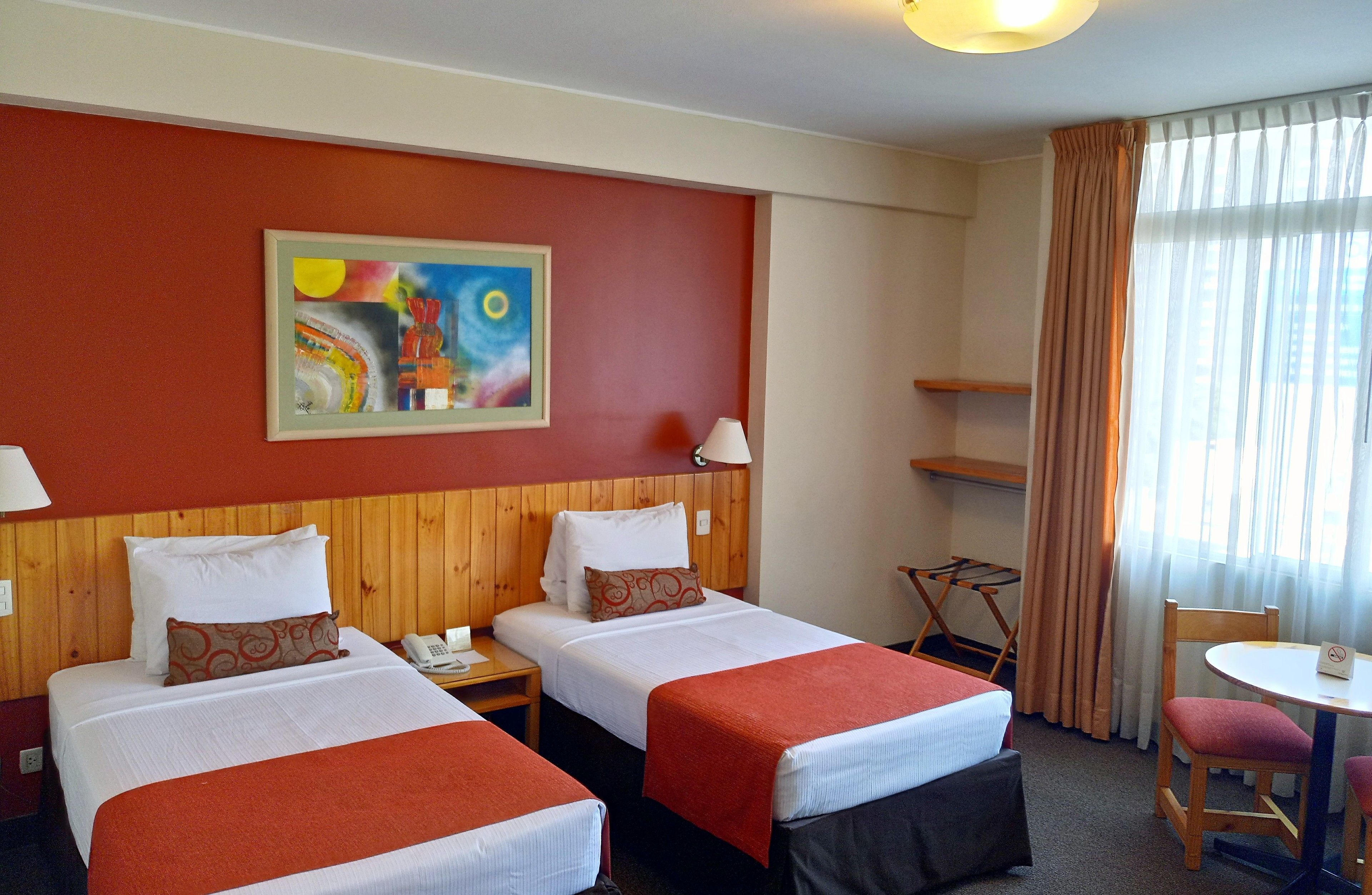 A hotel room with two beds, a painting on the wall, a desk with a lamp, and a chair.