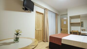 A hotel room with a bed, a round table with a vase of flowers, a television mounted on the wall, and a wooden door.