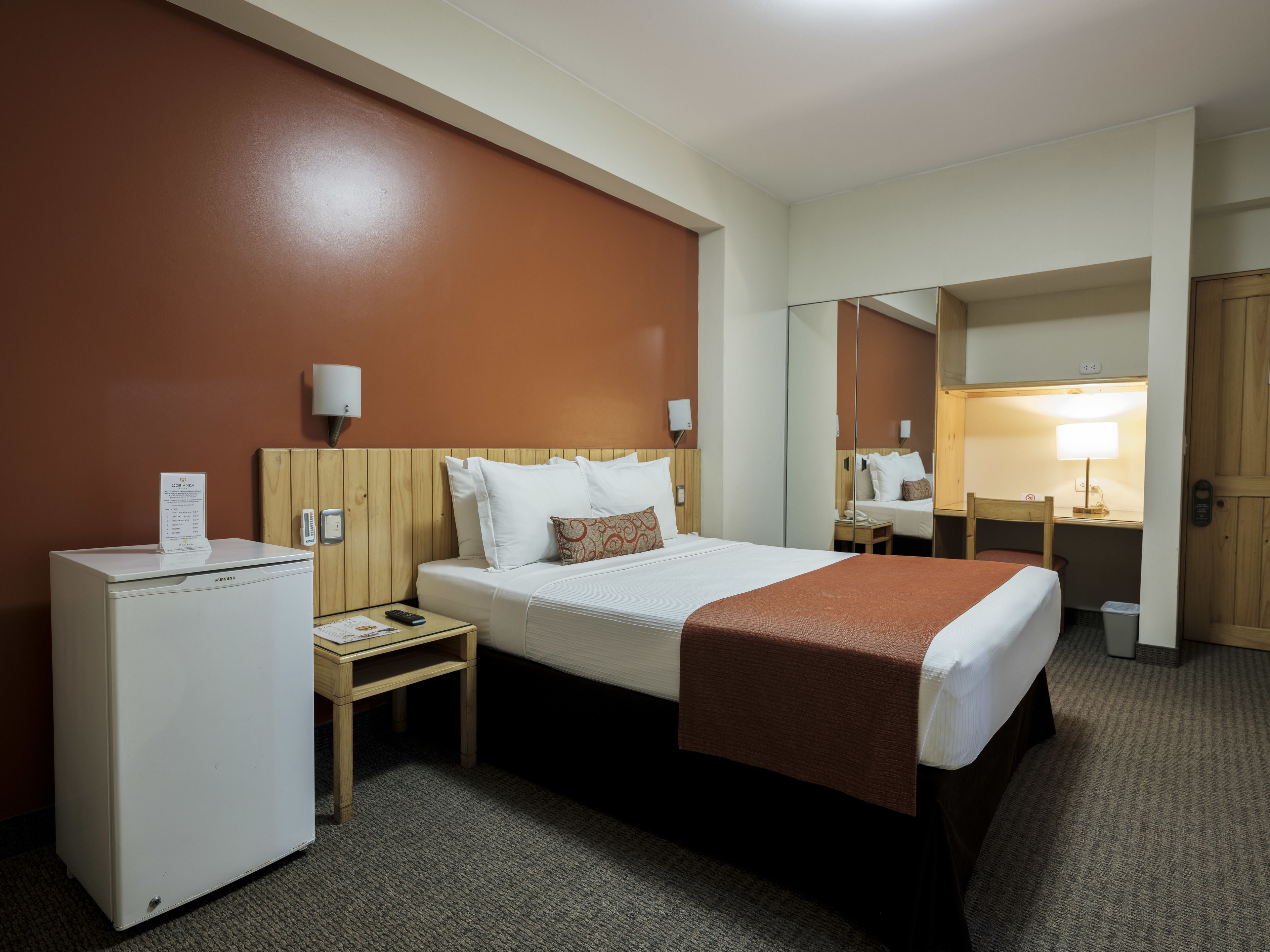 A hotel room with a bed, a desk, a chair, a refrigerator, and a mirror.