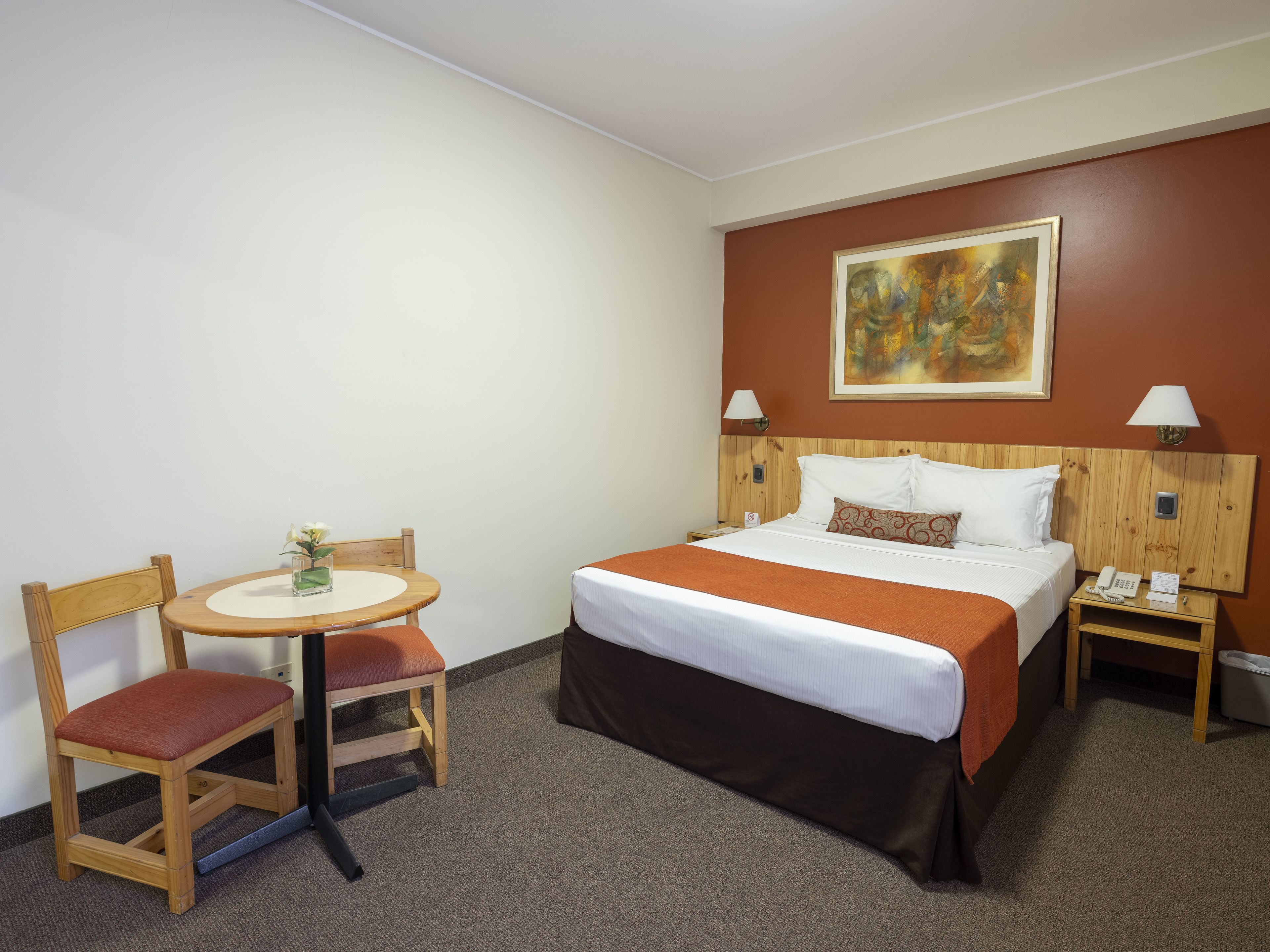 Ein Hotelzimmer mit einem Bett, einem kleinen Tisch mit Stuhl, einem Bild an der Wand und zwei Nachttischlampen.