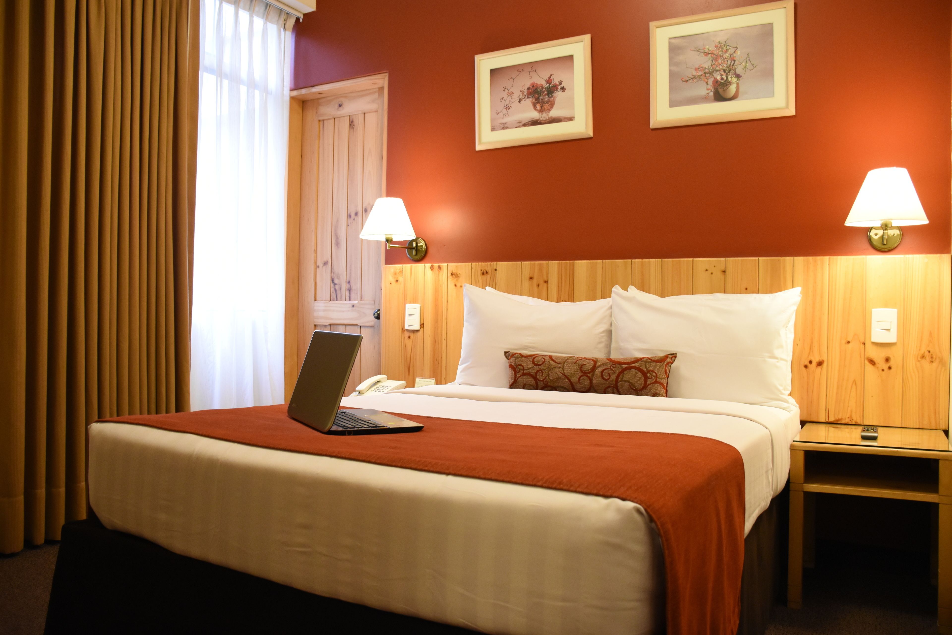 A hotel room with a bed, a laptop on the bed, two wall-mounted lamps, and two framed pictures on the wall.