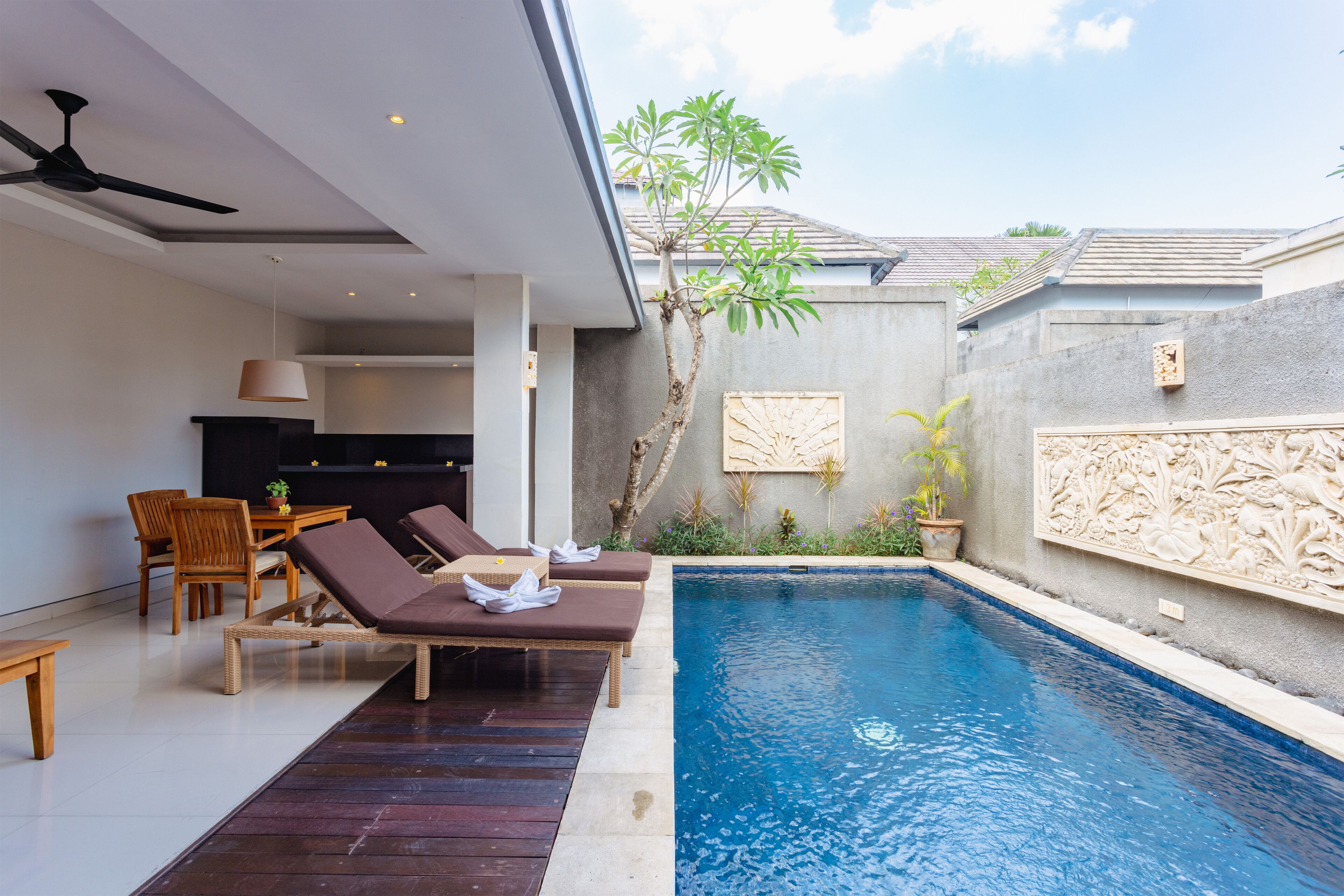 A modern outdoor pool area with lounge chairs, a dining table, and a ceiling fan.