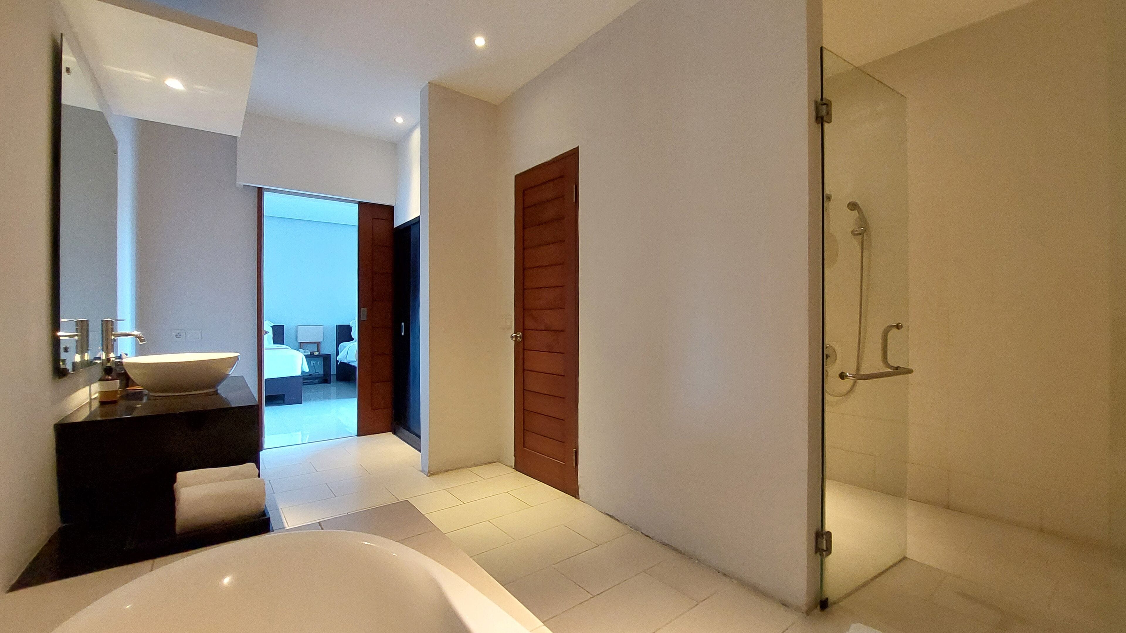 Un baño moderno con una bañera grande, un box de ducha de vidrio y una habitación visible al fondo.