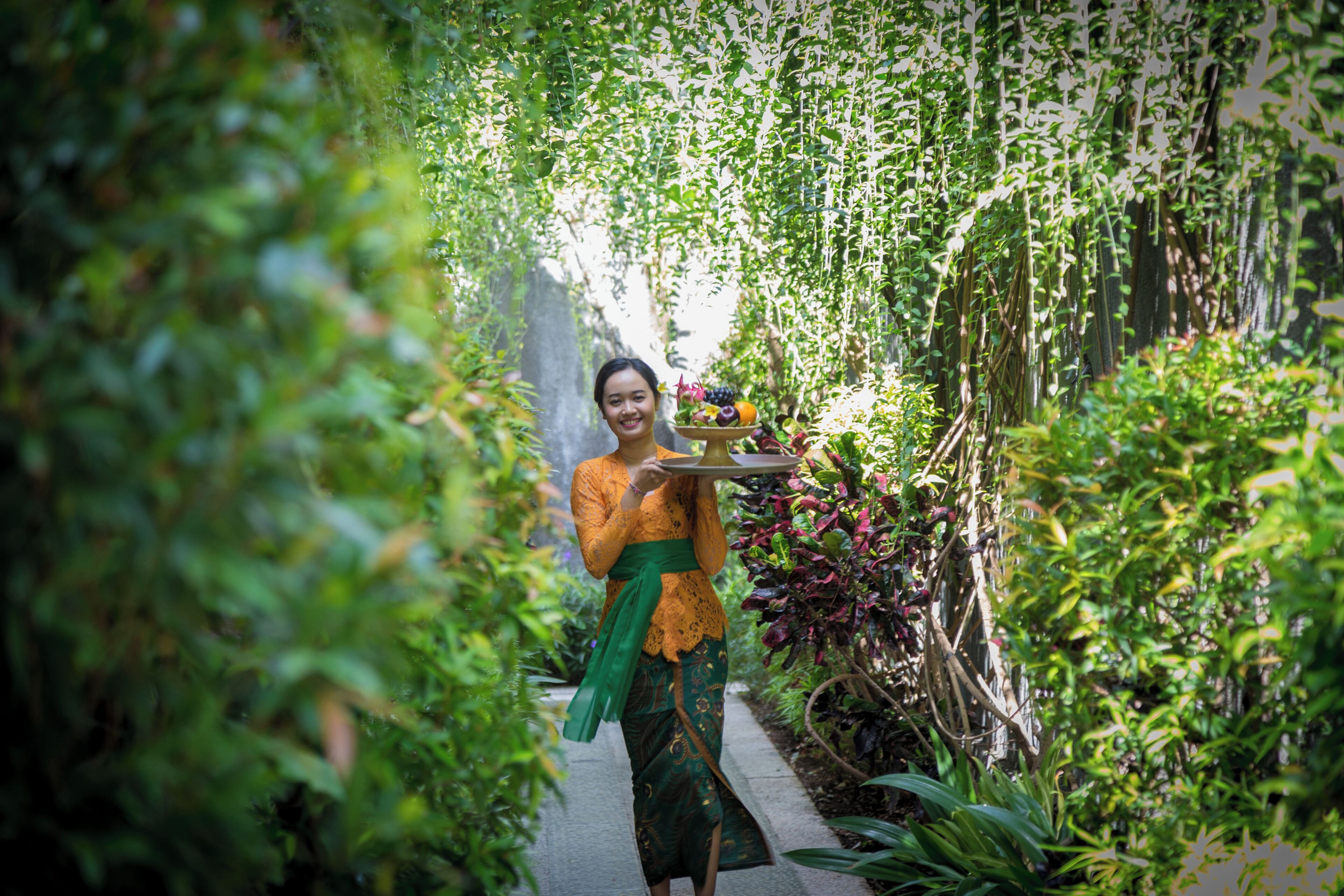 A person holding a tray with fruits and flowers, surrounded by lush greenery.
