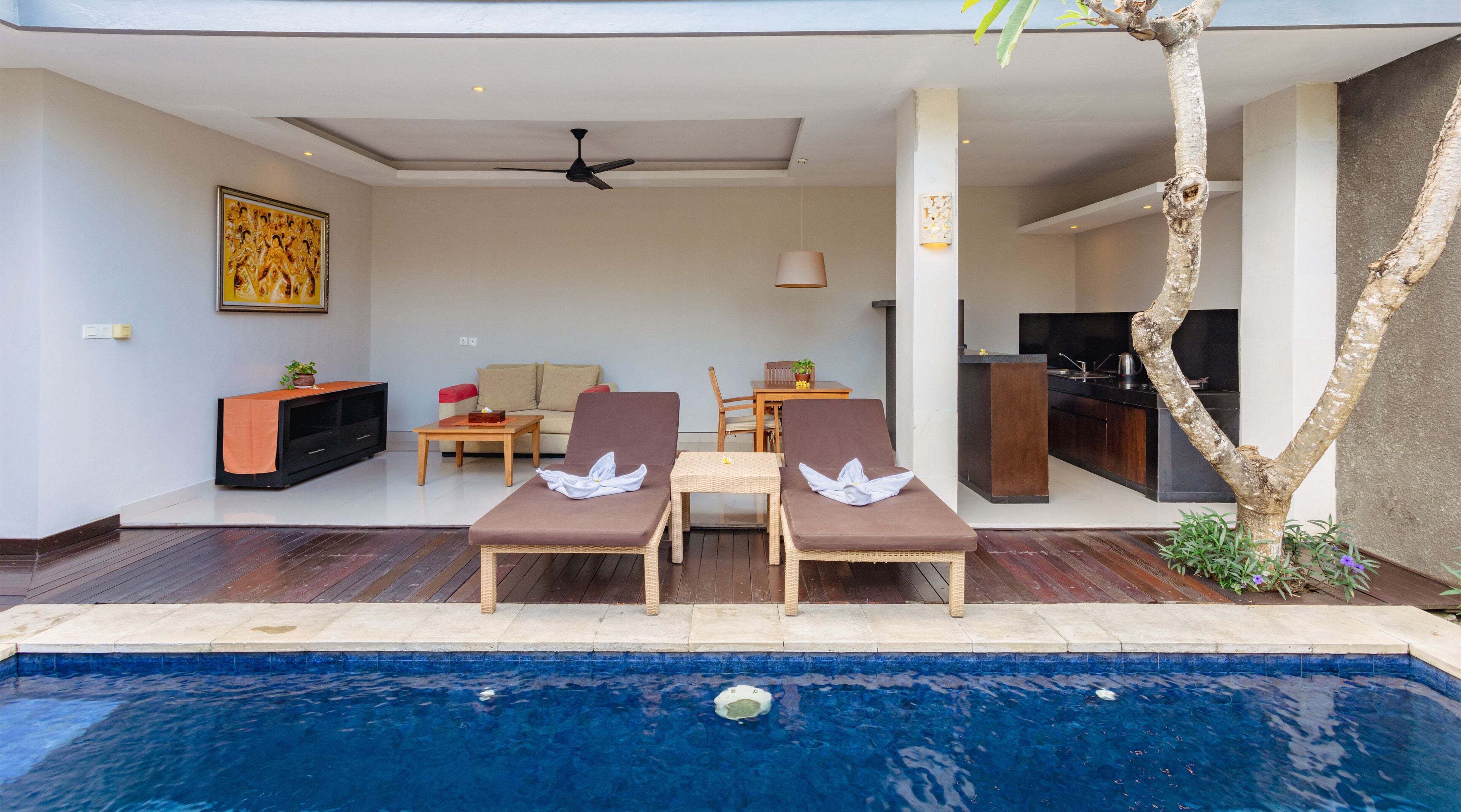 Un espace aménagé au bord de la piscine, avec des chaises longues, un ventilateur de plafond, une table à manger et une cuisine visible à travers une porte ouverte.
