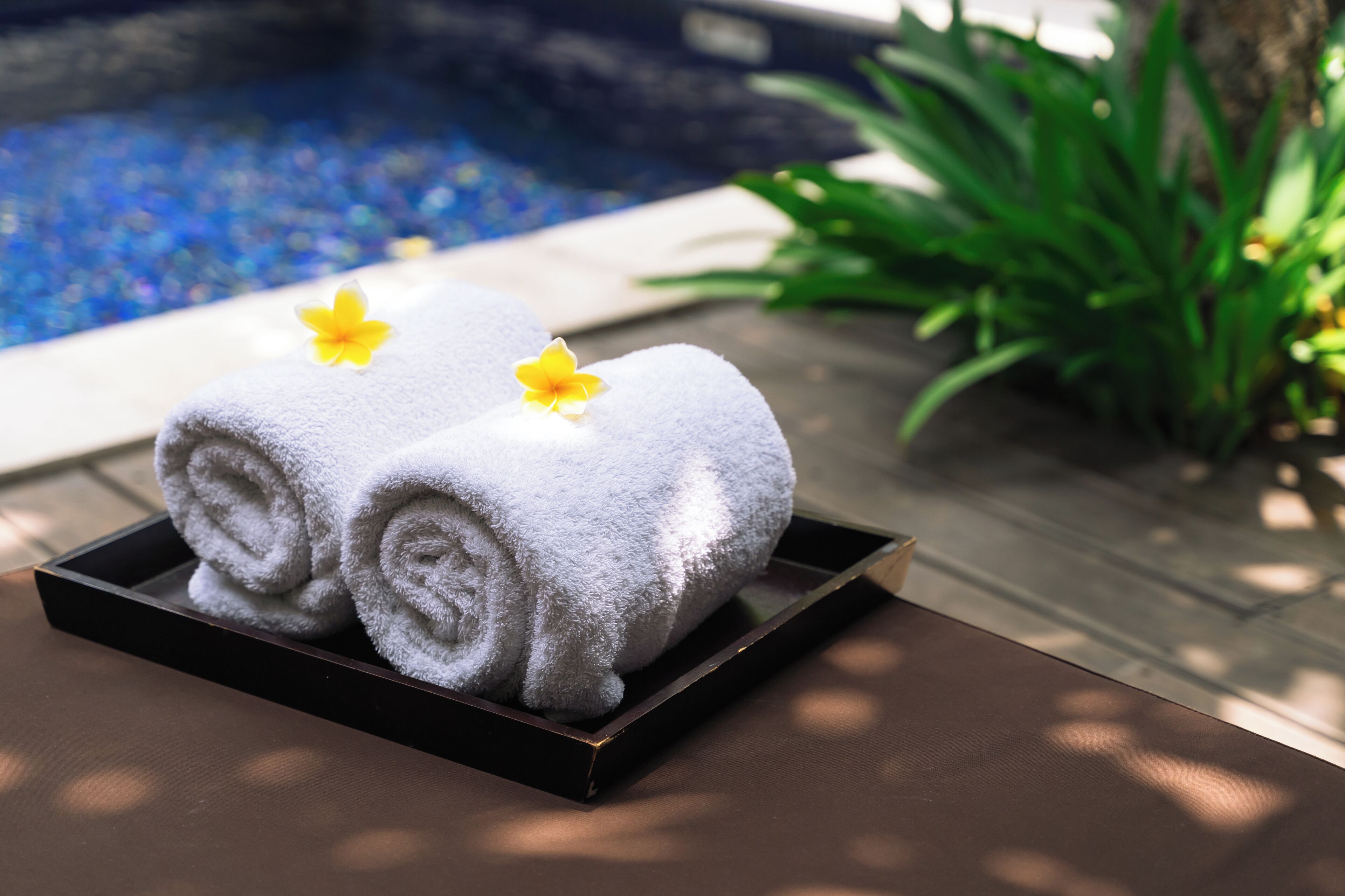 Un plateau avec deux serviettes enroulées et une fleur jaune, posé sur une table en bois près d’une piscine.