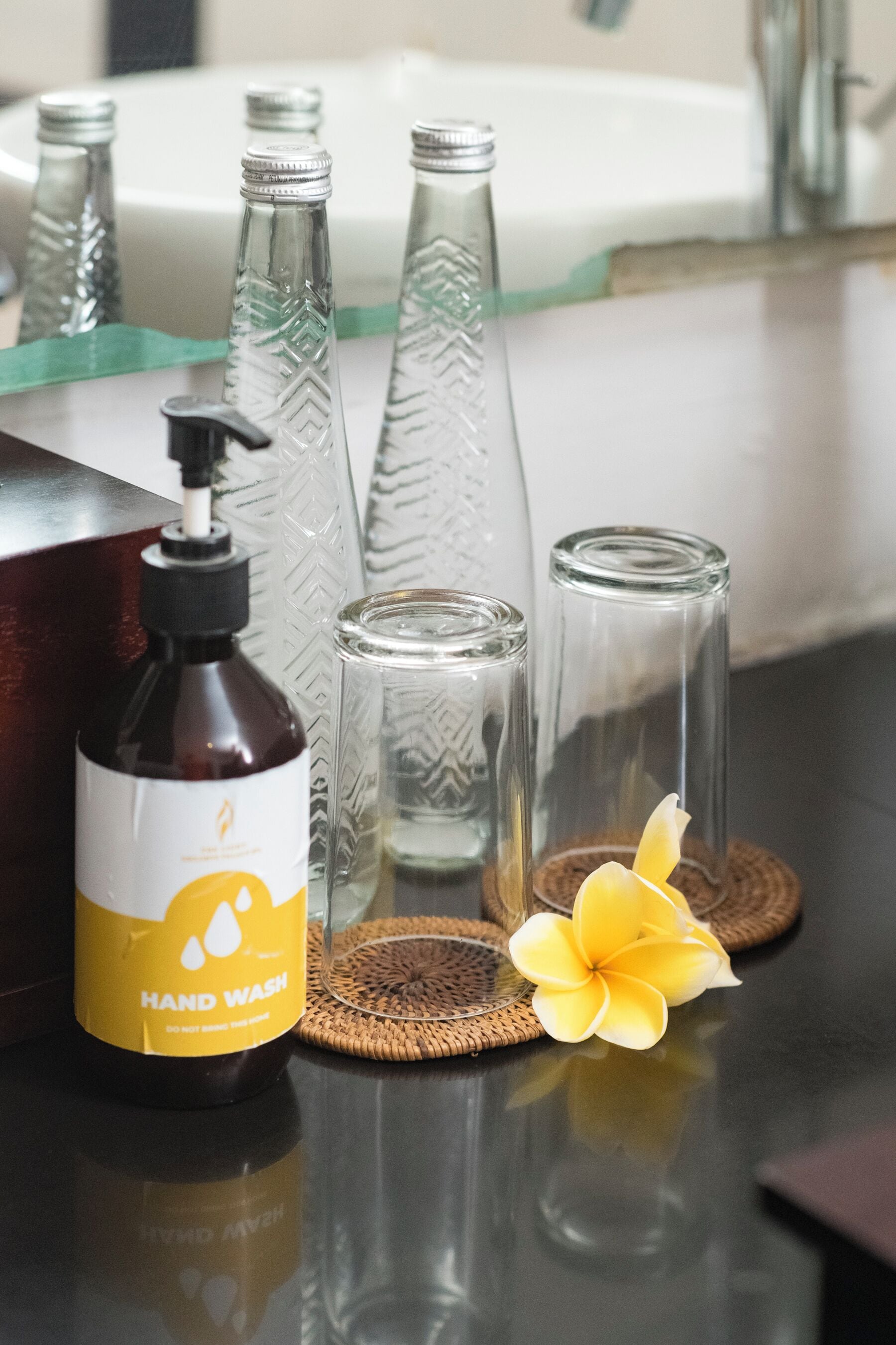 A hand wash station with a bottle, two glass bottles, and a yellow flower on a reflective surface.
