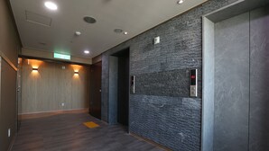 Un área de ascensor moderna con pared texturizada, panel de control del ascensor visible y una banda de pavimentación táctil en el suelo.
