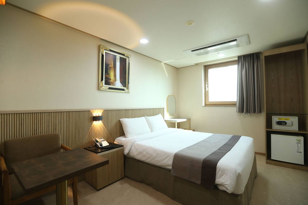A hotel room with a bed, a desk, a chair, a small refrigerator, a mirror, and a window with curtains.