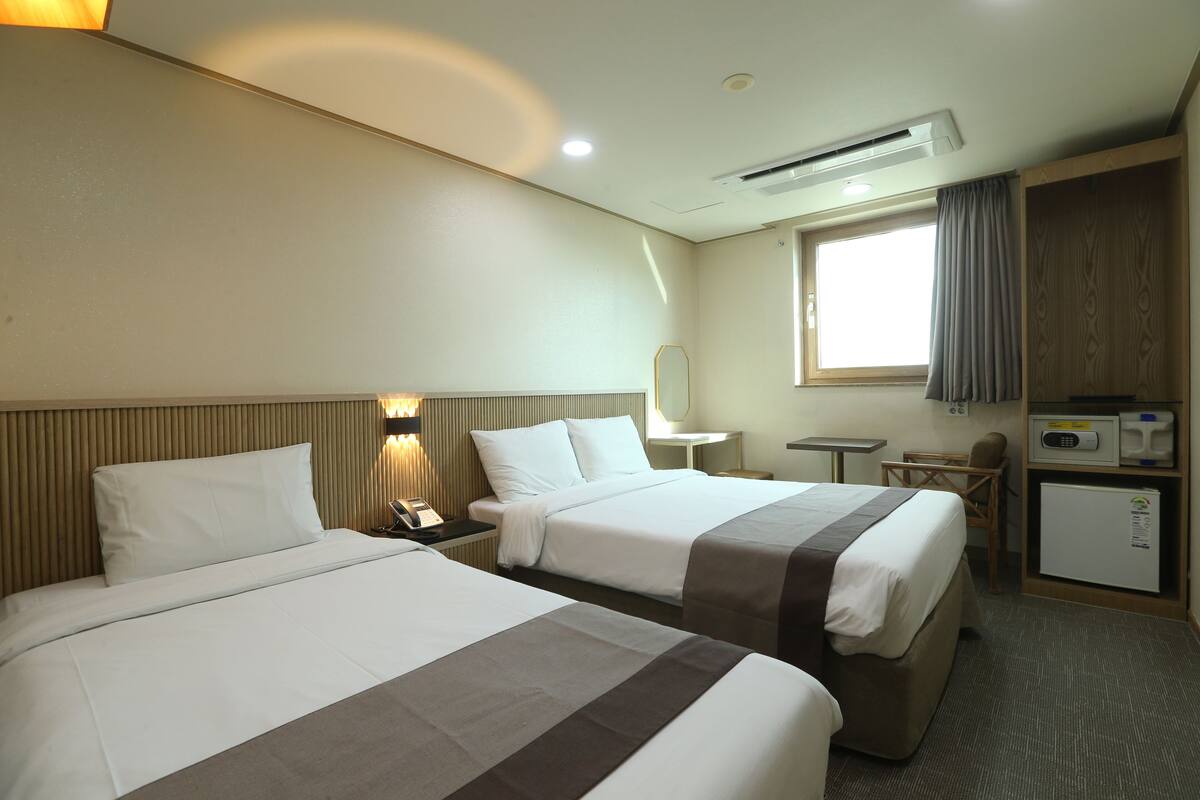 A hotel room with two beds, a desk, a chair, a small refrigerator, and a window with curtains.